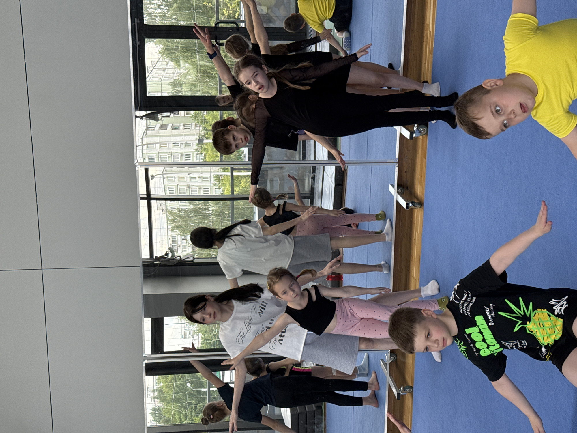 An indoor studio scene captures a group of children and a few adults engaged in what appears to be a dance, gymnastics, or stretching class. The floor is covered with a blue mat, and a large mirrored wall reflects the participants and the surrounding environment. Opposite the mirror, expansive windows offer a view of urban buildings and green trees, suggesting a city or suburban location, consistent with Khimki, Russia.
The participants, ranging in age from young children to pre-teens and teenagers, are mostly in poses with arms extended outwards or upwards, indicating a structured exercise or dance routine. Several individuals are focused on their reflections in the mirror or looking towards the camera.
Notable details include:
- A prominent girl in the center wearing a black long-sleeved leotard looks directly at the viewer.
- Another girl in the foreground wears a black tank top and pink polka-dotted shorts, with a focused expression.
- A young boy in a bright yellow t-shirt in the top right corner of the frame displays a playful, wide-eyed expression with his tongue slightly out.
- Another boy in the bottom right wears a black t-shirt adorned with a green pineapple graphic and visible text: "POSITIVE MND / POSITIVE VRES POSITIVE LIVE," "cool itamin," and "Summer Holiday."
- An adult in a white t-shirt has text printed on it that reads: "The Light will Eve ntua."
- The bright natural light filtering through the windows indicates it is daytime.
The image conveys an energetic atmosphere of learning and physical activity within a dedicated studio space.