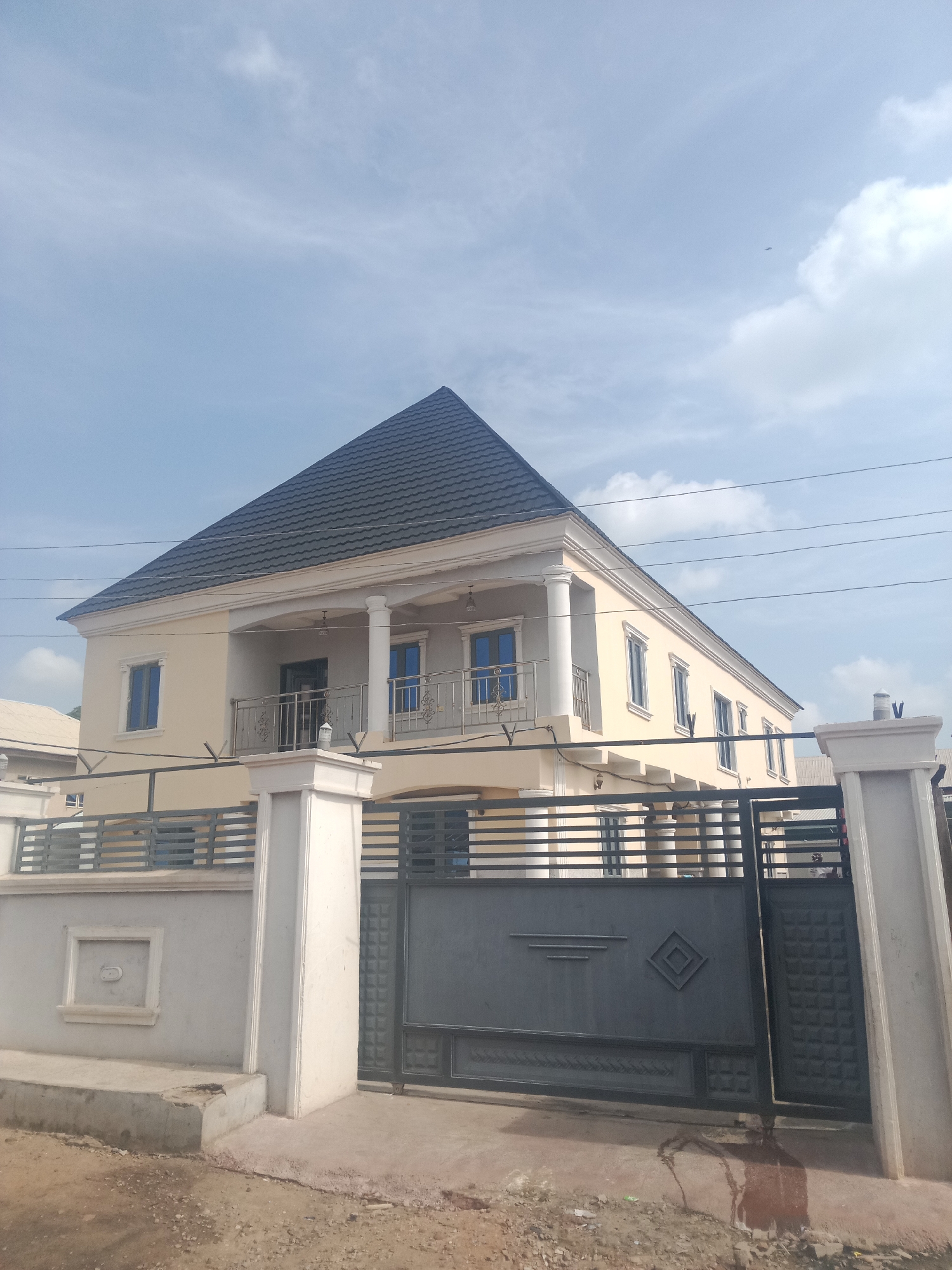 A two-story house with a dark, pyramid-shaped roof and light yellow walls is visible behind a grey metal gate and a white fence. The house has several blue-tinted windows and a balcony on the second floor. The gate is closed, and the fence is made of horizontal bars with decorative elements. The ground in front of the gate is a mix of concrete and dirt. The sky is light blue with some white clouds. This scene is located in Lokoja, Nigeria.