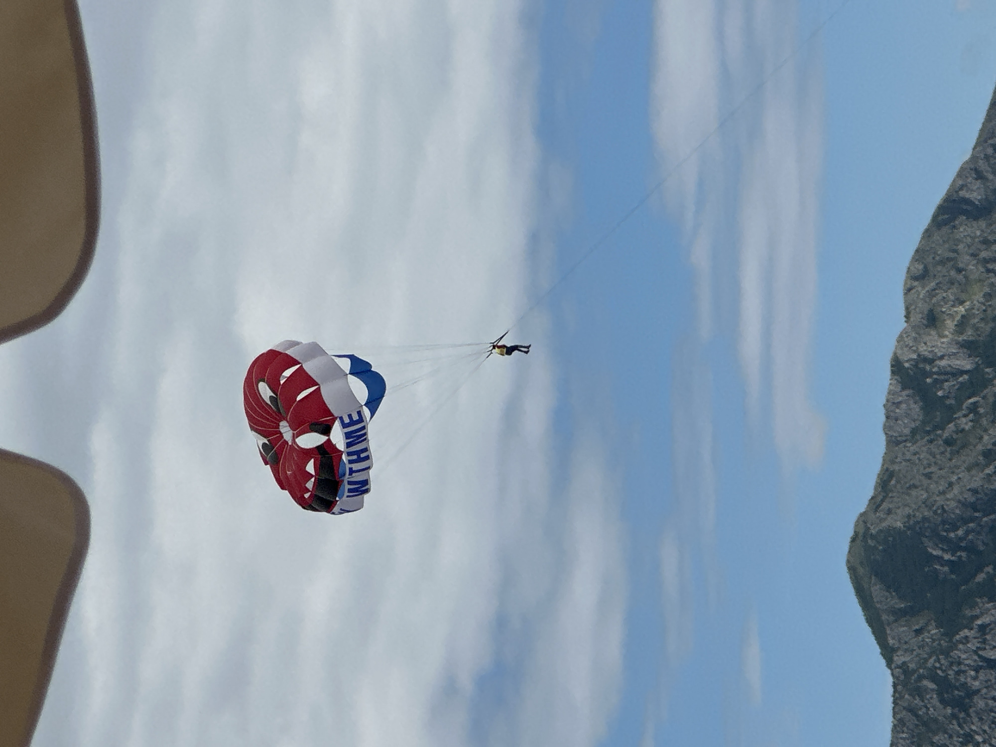 An outdoor, high-angle shot captures a person parasailing against a bright, partly cloudy sky, likely over water near Budva, Montenegro. The central subject is a colorful parasail, predominantly red, white, and blue, with parts of a word, "WINTHME", visible on its surface. Below the parasail, a single person is suspended in a harness, facing away from the camera, enjoying the airborne activity. A thin tow line extends upwards and to the right from the parasailer, indicating they are being pulled by an unseen boat.
The background features a vast expanse of light blue sky dotted with scattered white clouds. To the far right, a large, dark grey, rocky mountain or cliff face is visible, suggesting a coastal or mountainous setting. In the top-left corner, a partial view of a light brown, curved object, possibly a hat or part of the viewing platform (e.g., a boat canopy), frames the scene. The lighting indicates it is daytime, with clear visibility. The image conveys the thrill and serene beauty of parasailing in a scenic location.