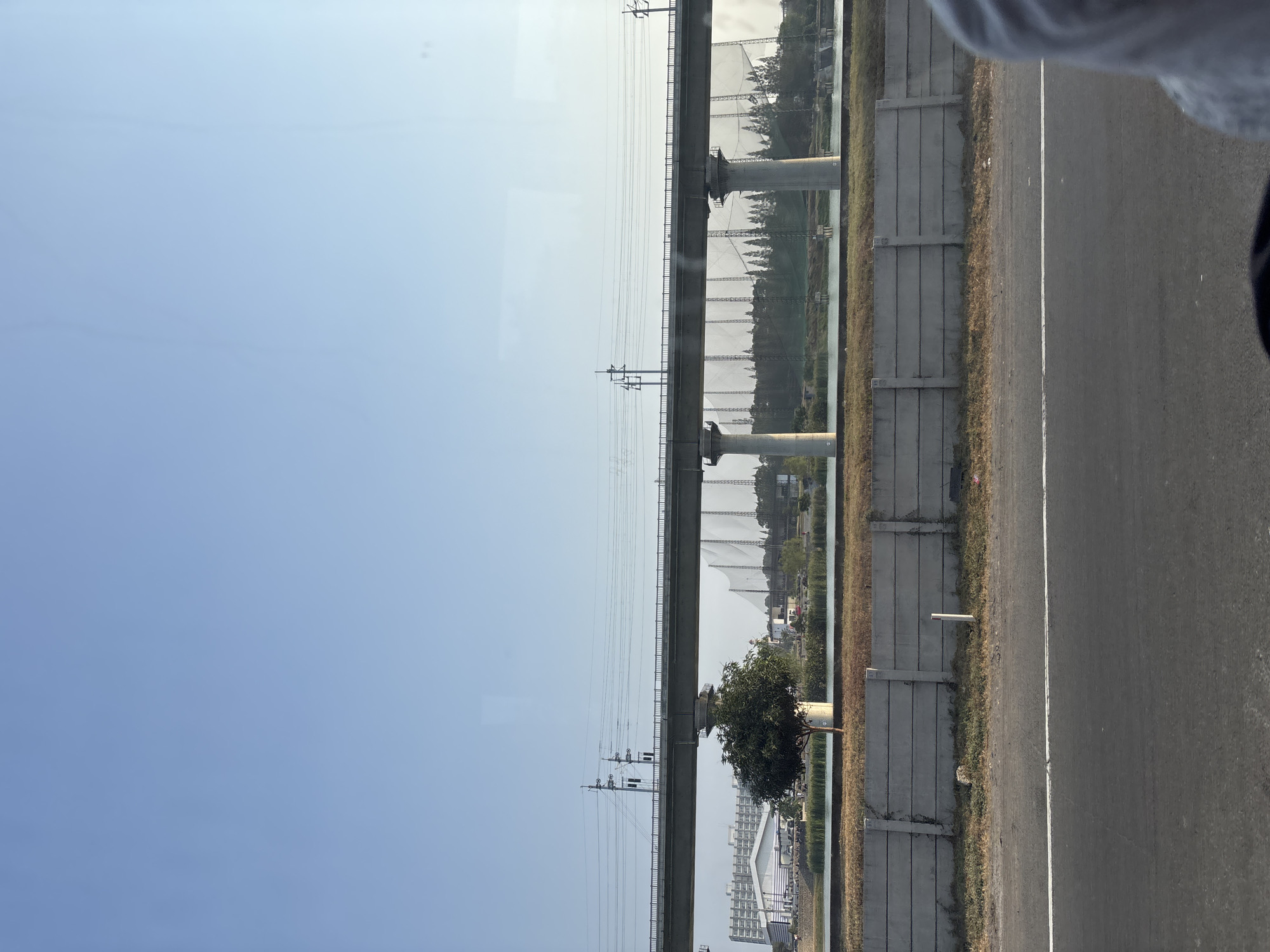 A wide shot captures a concrete overpass with utility poles and wires overhead, spanning across a body of water. A lone tree stands near the water's edge. In the background, a golf driving range with tall nets is visible, along with a low-rise building and a cluster of trees. A concrete barrier runs along the foreground, separating the road from the embankment. The sky is clear and blue.