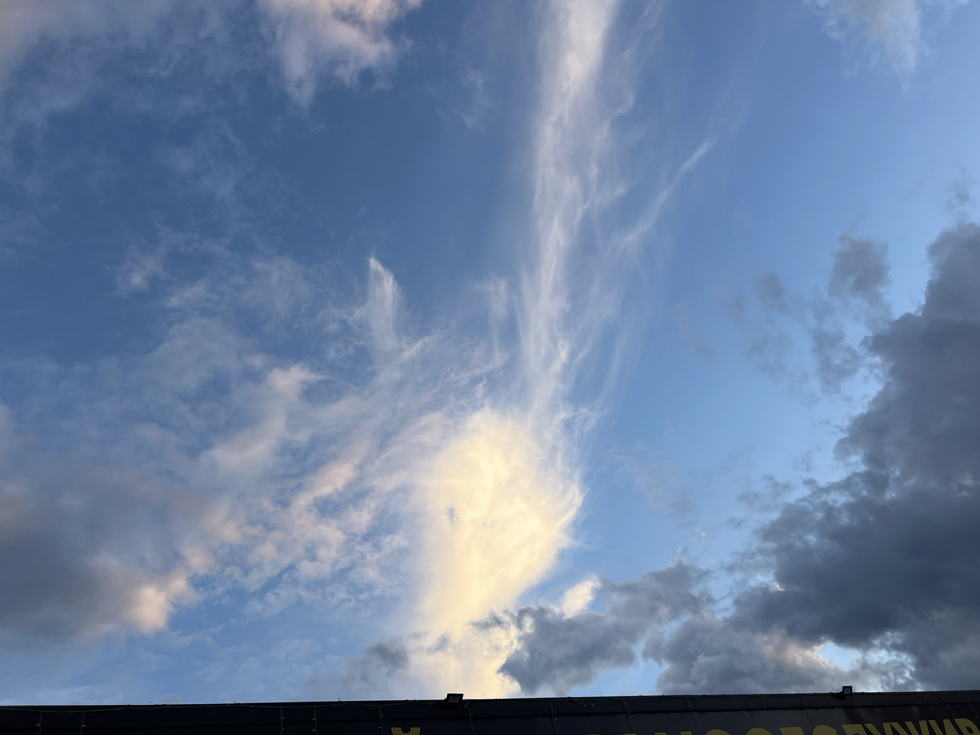 The image shows a view of the sky with a mix of cirrus and cumulus clouds. The clouds range in color from bright white to dark gray, indicating varying levels of sunlight and moisture. The sky is blue in some areas. At the bottom of the frame, a portion of a dark-colored structure is visible with some text that includes the name "Chaikovsky" in Cyrillic letters, suggesting that the image was taken in or near the city of Chaikovsky, Russia.