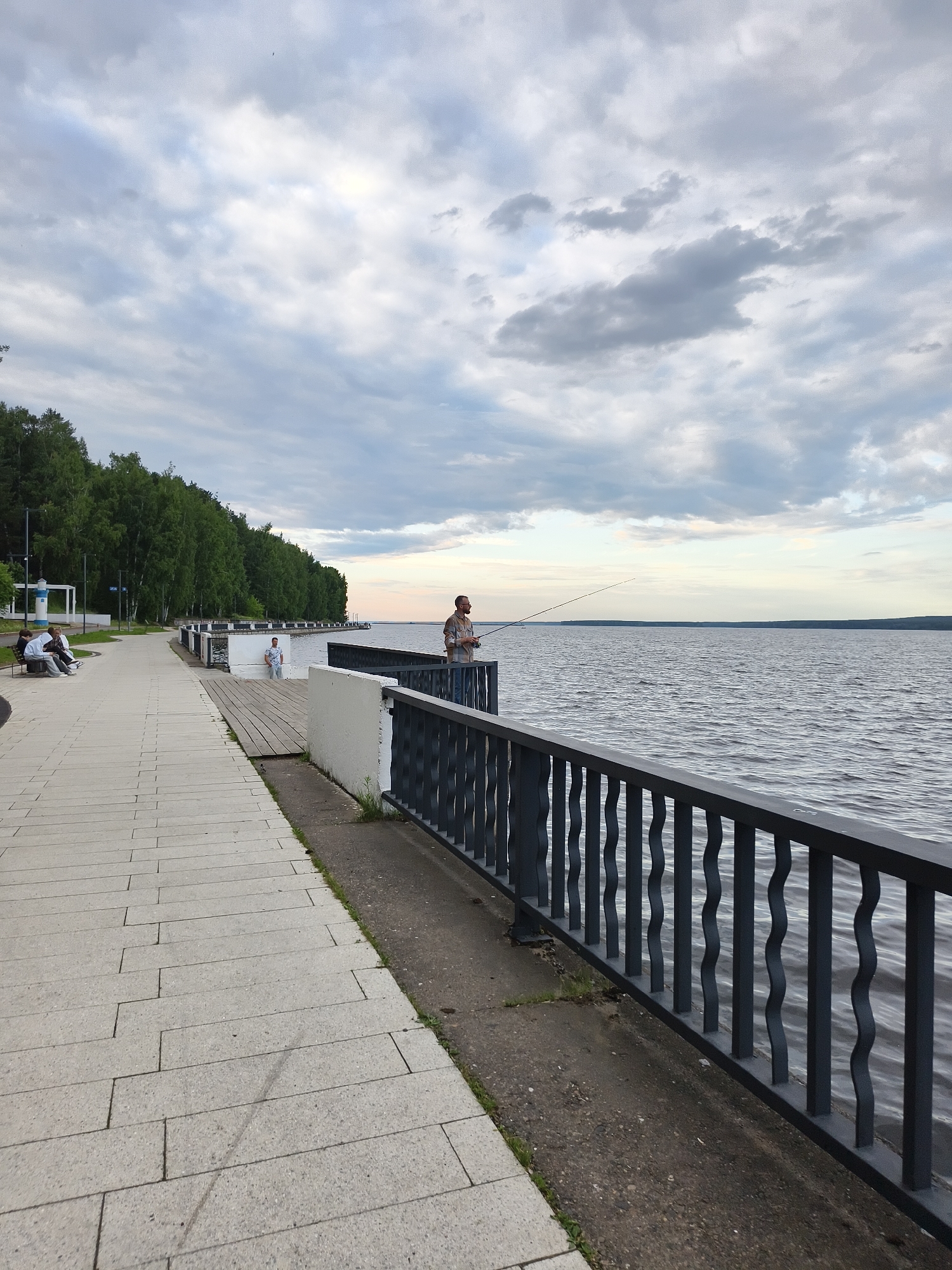 The image depicts a wide paved walkway along a body of water, likely the Kama River, in the city of Chaikovsky, Russia. A dark metal railing with a wavy pattern separates the walkway from the water. On the right side of the walkway, a man is fishing with a rod. Further along the walkway to the left, two people are seated on a bench, and another person stands near a white structure that may be a small lighthouse or information kiosk. The background features a dense forest of green trees bordering the water's edge. The sky is overcast with scattered clouds. The scene appears to be a public promenade or embankment.