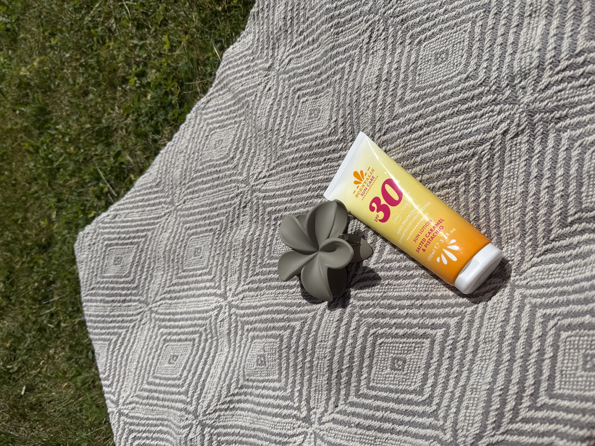 The image shows a tube of Scentalis Sun Care SPF 30 sun lotion resting on a gray and white patterned blanket, next to a gray flower-shaped hair clip. The blanket is partially placed on green grass, suggesting an outdoor setting. The sun lotion tube is primarily yellow and white with the "SCENTALIS SUN CARE" branding visible. The blanket features a geometric pattern composed of squares and diamonds.
