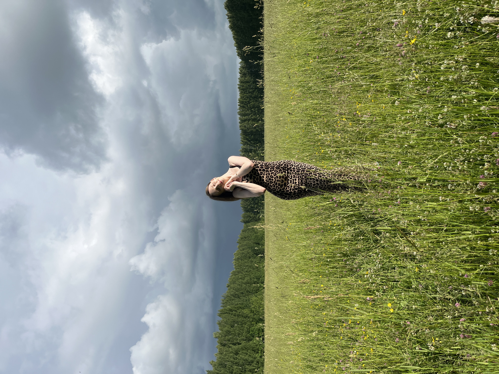 A full-body shot of a young woman in a leopard-print maxi dress stands in a field of tall green grass and wildflowers. The woman is positioned in the center of the frame, with her head tilted back and eyes closed, her hands clasped together near her face in a pose of serene contemplation. The field is lush with a variety of green grasses and small white and pink flowers, with a few yellow wildflowers interspersed. In the background, a dense line of dark green trees stretches across the horizon, forming a stark contrast against a dramatic sky filled with heavy, dark gray clouds, suggesting an impending storm. The overall lighting is subdued due to the cloud cover. The location is the outskirts of Большой Гондыр, Russia.