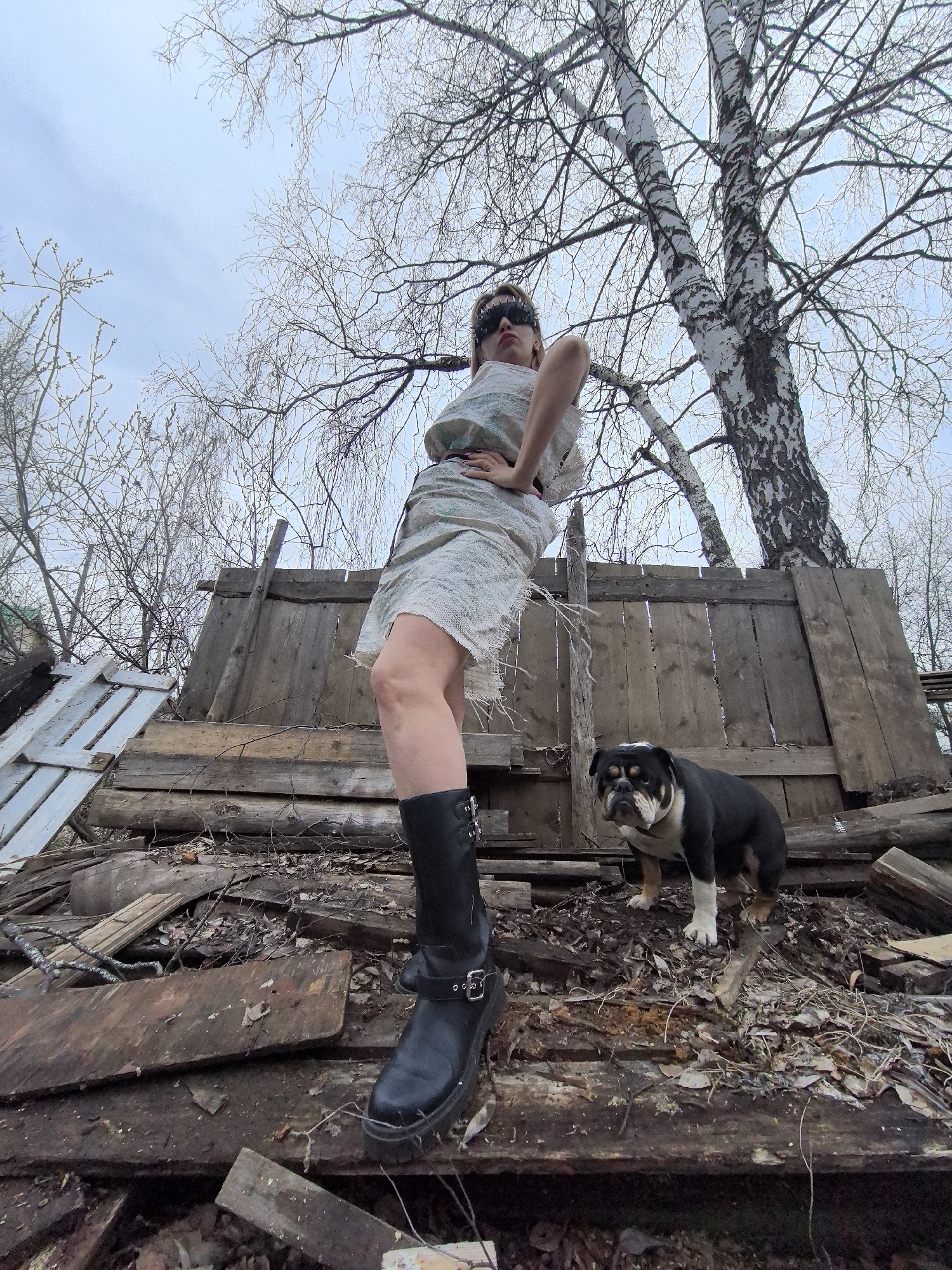 A low-angle, full-body outdoor shot depicts a woman and a bulldog standing atop a pile of wooden debris in Noviy, Russia. The woman, positioned centrally, has her hands on her hips in a confident pose, looking down towards the camera. She wears a light-colored, possibly distressed, knee-length dress or tunic, black chunky boots with buckles, and dark, spiked sunglasses or a visor. Her lips are painted red. To her right, a black and white bulldog with tan markings stands on the wooden planks, looking intently at the viewer.
The setting appears rustic and somewhat desolate, with a jumble of wooden planks, possibly from a fence or dilapidated structure, forming the foreground and the elevated platform for the subjects. Above them, bare-branched trees, including distinctive white-barked birches, stretch upwards against a light, overcast sky. The absence of leaves suggests late winter or early spring. The low camera angle emphasizes the subjects against the expanse of the sky and trees, giving the woman an imposing presence. The time of day appears to be daytime under cloudy conditions. There is no visible text in the image.