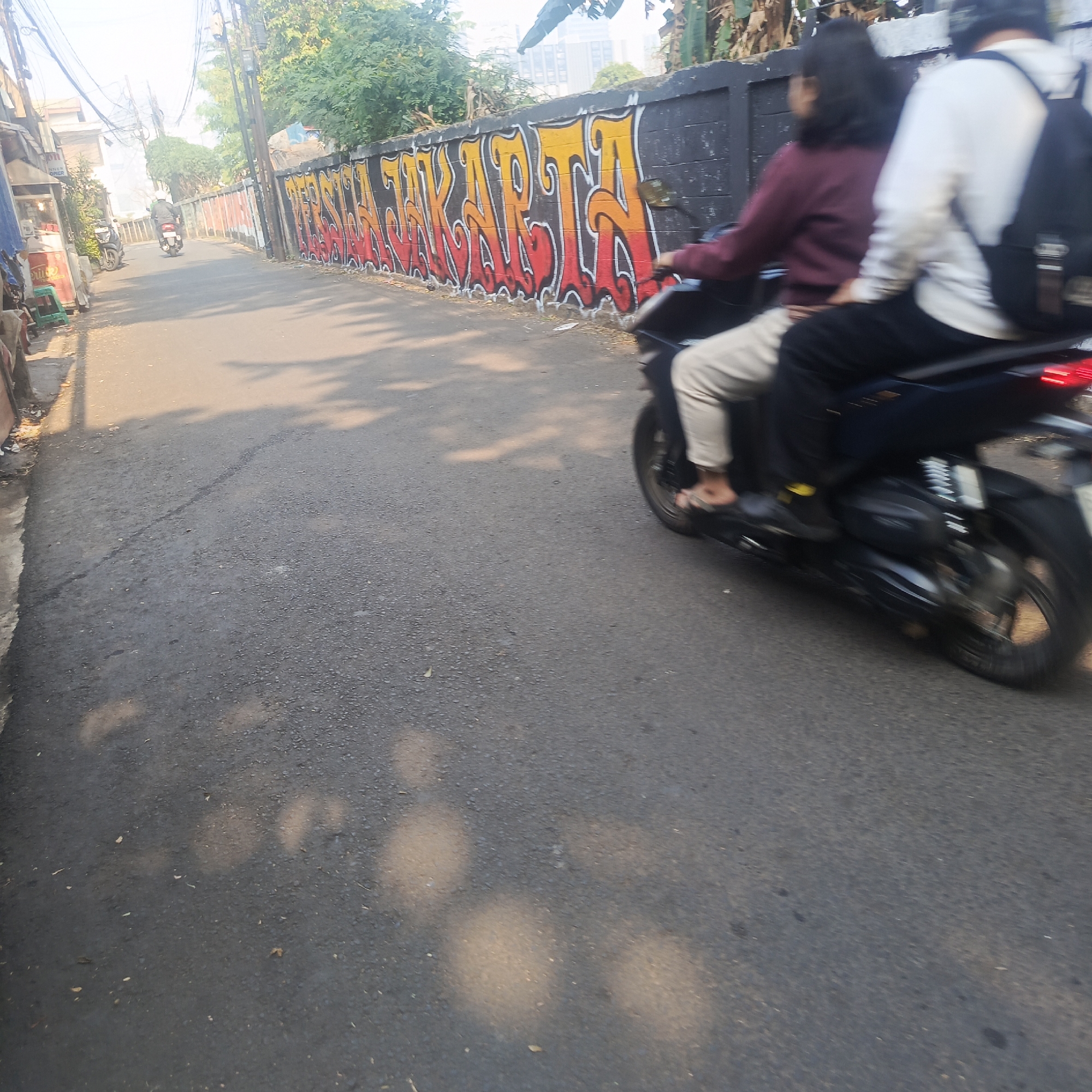 The image captures a street scene in South Jakarta, Indonesia, characterized by a road surface with visible tire marks and scattered debris. On the right side of the road, a black motorcycle is in motion, with two blurred figures on it. The rear passenger, wearing a white long-sleeved shirt and a backpack, is prominently visible. The front passenger, wearing a maroon long-sleeved shirt and light-colored pants, is also discernible. The motorcycle has a red taillight that is illuminated.
Adjacent to the road, on the right, is a black wall adorned with vibrant graffiti in shades of yellow and red, featuring stylized lettering. On the left side of the road, there are buildings, including a shop with a green plastic stool and a sign that reads "NINTY SNER". A few trees are present, with foliage visible. The overall lighting suggests it is daytime, with shadows cast across the road, indicating a sunny environment. The motion blur of the motorcycle and its riders suggests movement down the street.