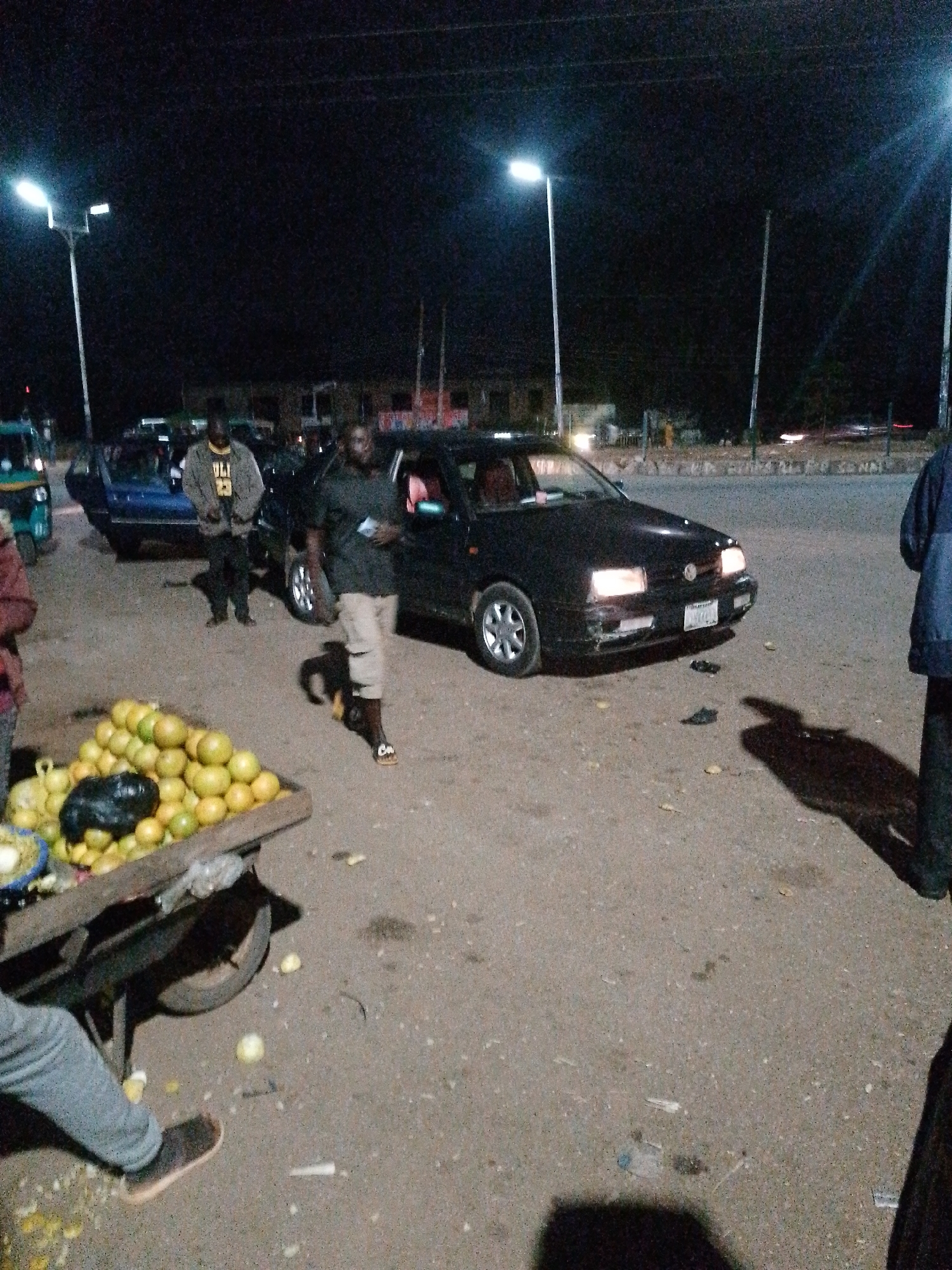 The image depicts a nocturnal street scene in Bukuru, Nigeria, illuminated by streetlights and vehicle headlights. In the foreground, a wooden cart is laden with a large pile of oranges and some black plastic bags. Next to the cart, a person wearing grey pants and black shoes stands with their left leg bent. To the right of the cart, a dark-colored car is parked, its headlights on. A man in a dark shirt and light-colored shorts walks past the car, holding a small object in his right hand. Another man, wearing a jacket over a t-shirt, stands near the car. Further in the background, other vehicles and buildings are visible, as well as streetlights. The ground is a mixture of asphalt and gravel.