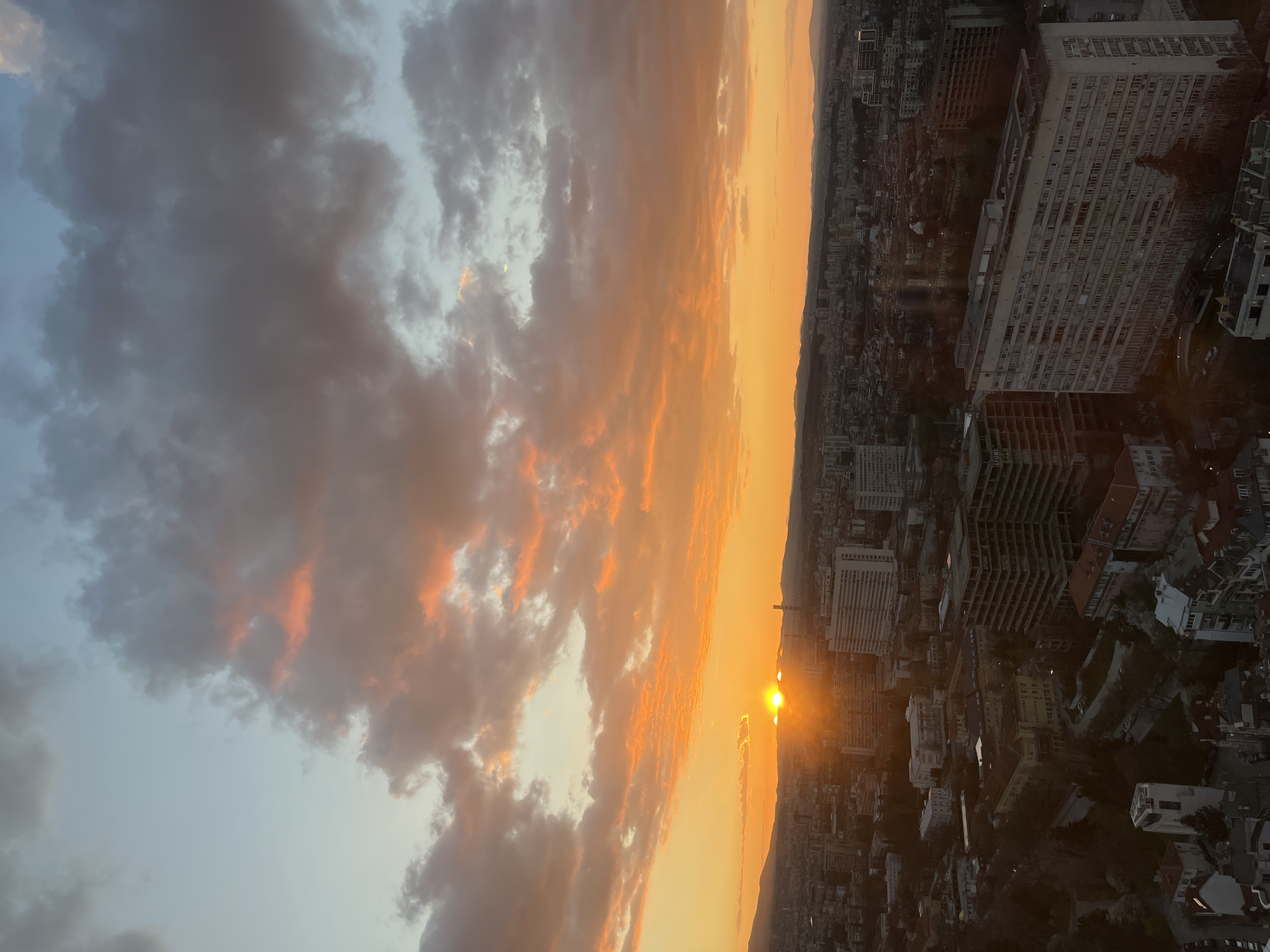 The image depicts a sunset over Sofia, Bulgaria. The sky is filled with clouds illuminated by the setting sun, casting an orange glow across the scene. Below, the city sprawls, with buildings of various sizes and styles visible. The architecture suggests a mix of older and more modern structures. The sun is positioned near the horizon, shining through a gap in the cityscape.