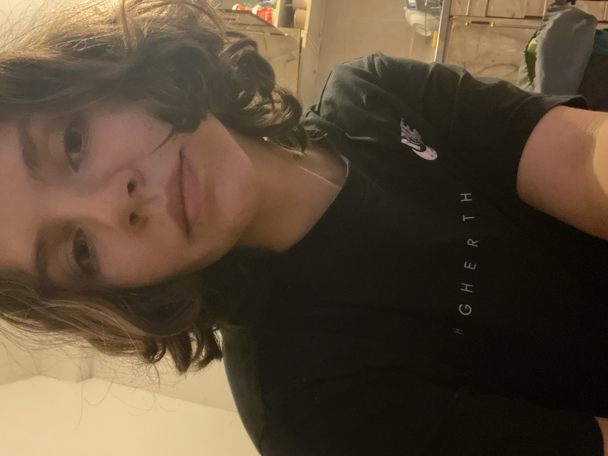 A medium shot depicts a young individual with light skin and curly brown hair wearing a black T-shirt with the word "HIGHERTH" printed vertically on the right side and a small white Nike logo on the upper left. The individual is looking towards the camera with a slight smile. The background appears to be an indoor setting, possibly a room in a house or apartment, with shelves visible on the wall behind the individual. The exact location in the city of Budva, Montenegro, cannot be determined from the image.