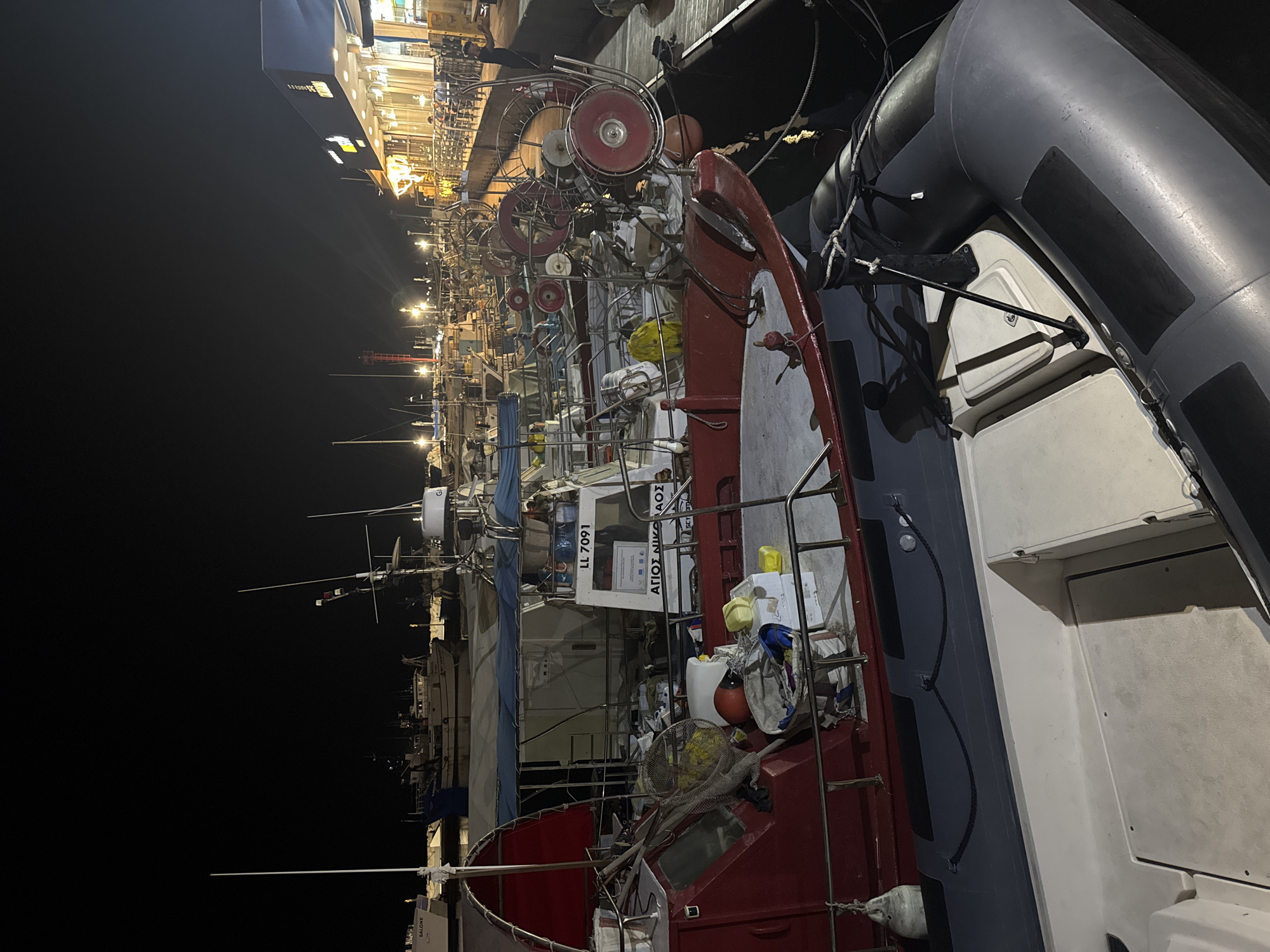 A nighttime scene at a harbor in Limassol, Cyprus, features a row of docked fishing boats and a marina walkway with illuminated buildings. In the foreground, a large inflatable boat is moored next to a red fishing vessel. The fishing boat has various equipment onboard, including nets, buoys, and storage containers. The vessel is identified by the name "ΑΓΙΟΣ ΝΙΚΟΛΑΟΣ" and the registration "LL 7091". Further down the dock, other fishing boats are visible, some with lights on. The marina walkway is lined with tables and chairs, suggesting a waterfront dining or social area. The background shows more boats and the dark expanse of the sea. The lighting is artificial, casting long shadows and creating a slightly dramatic atmosphere. There are no discernible people in active interaction, but a few figures can be seen on the marina walkway in the distance.