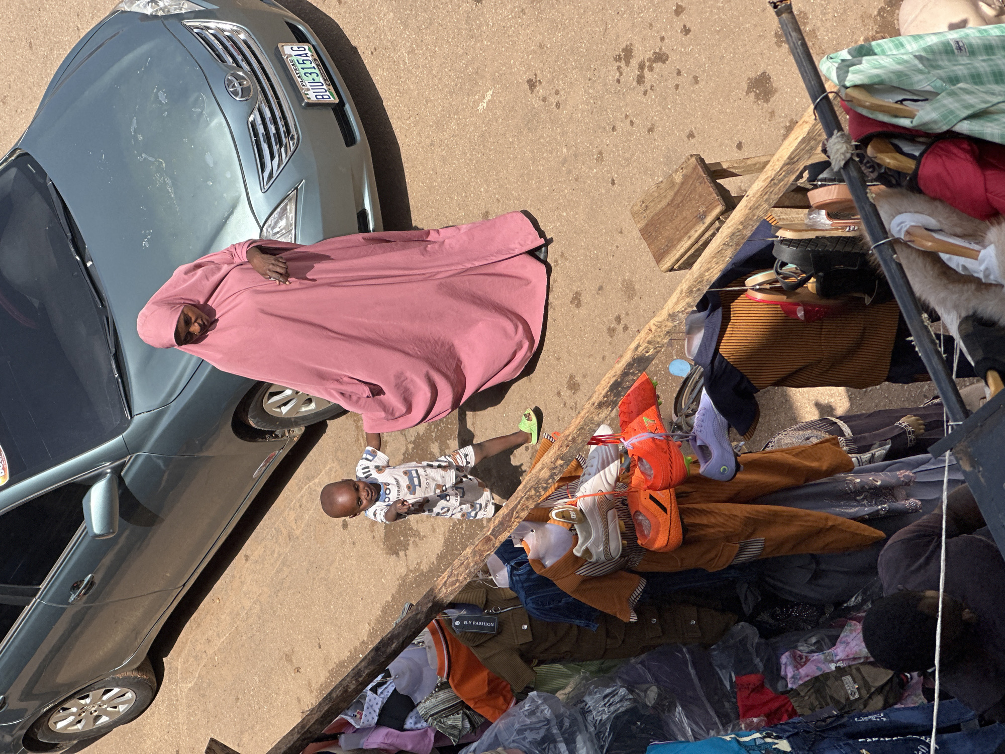 In Bukuru, Nigeria, a woman in a pink hijab stands next to a parked teal sedan, her gaze directed downwards. Beside her, a toddler in a striped outfit points towards the foreground, where a market stall displays various garments and footwear. Among the items are orange soccer cleats, purple sneakers, and an assortment of clothing items, including pants, shirts, and dresses. The ground, a dusty, reddish-brown surface, is speckled with dark splotches, possibly from spilled liquids or mud. A wooden stool sits near the stall, and further back, more market goods are visible, suggesting a bustling commercial area. The sun casts sharp shadows, indicating a bright, daytime scene.