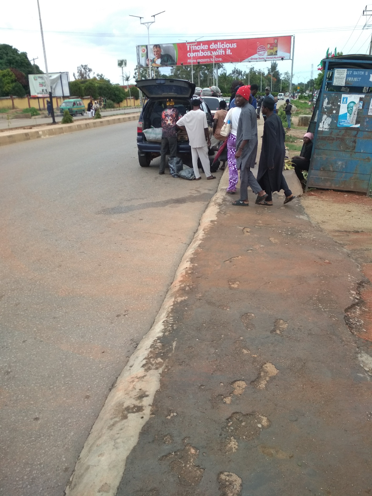 This image depicts a bustling street scene in Jos, Nigeria, under an overcast daytime sky. In the midground, a dark-colored SUV is parked on the roadside with its tailgate open. Several individuals are gathered around the vehicle, appearing to be loading or unloading items from the trunk. People are dressed in a mix of traditional and casual attire; one person wears a distinctive red cap, while others are in white, grey, and black garments.
To the far right, a blue, box-like structure stands on the sidewalk, with a person sitting beside it. This structure features posters, one of which displays a man's face and text including "HABILITERS URGENTLY," "17 BATCH B," "GLOBAL STAR PROJECT," and a phone number "20184707917."
In the background, along the road, a green mini-bus is visible further down the street. Overhead, large billboards are prominent. A red billboard advertises "Nnake delicious combos with it. More reasons to love," highlighting "Pure Protein" for "Great Taste, Good Nutrition." Another green billboard on the left partially displays "TEAL" and "EL BANU ANK." The foreground shows a worn asphalt road alongside a concrete curb, bordering a patchy and uneven dirt/gravel sidewalk. The overall scene suggests a busy urban or semi-urban transit point.