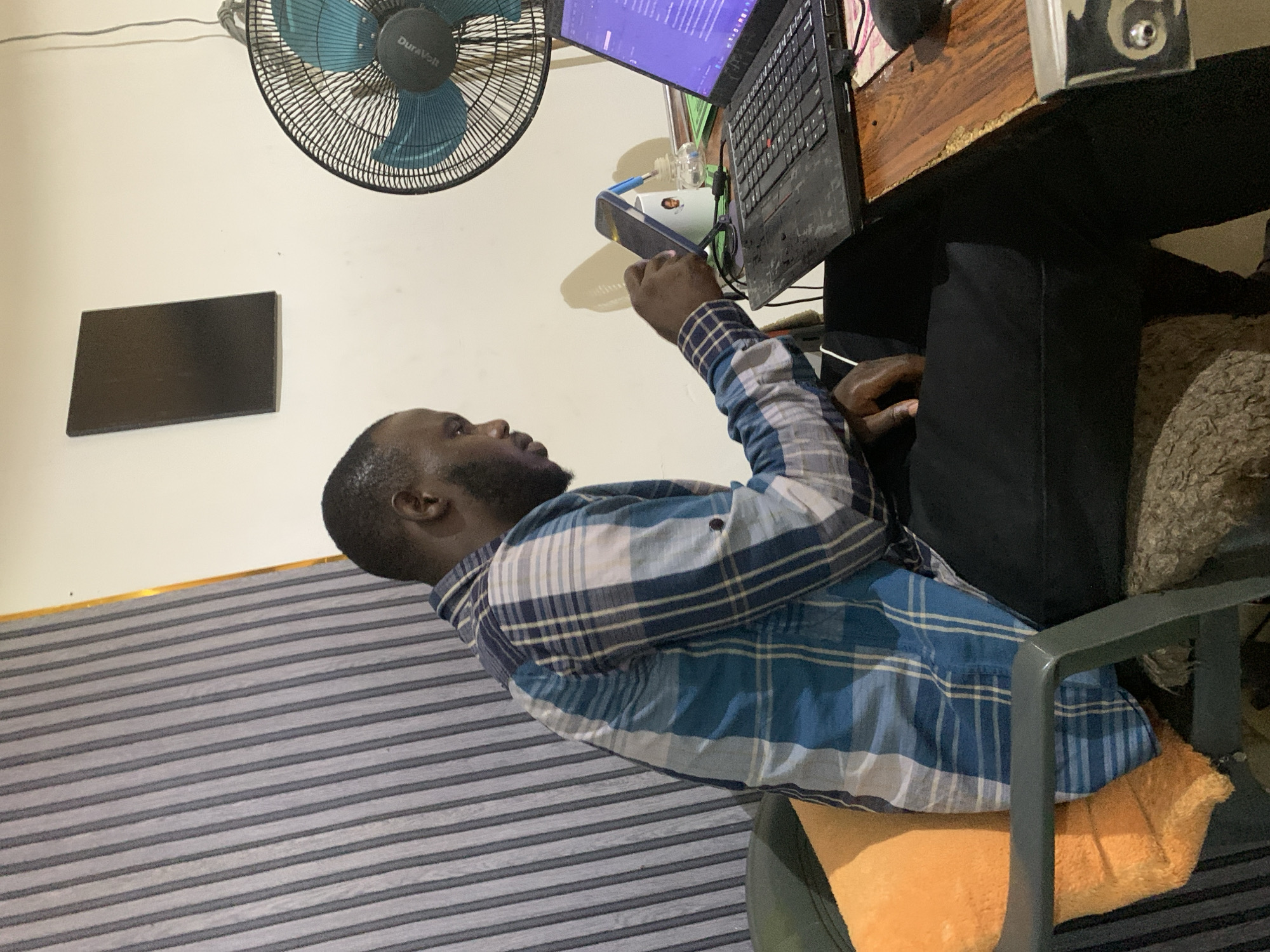 A man is seated at a desk in an interior space. He is Black, with a shaved head and a beard. He is wearing a blue and white plaid shirt and black pants. He is holding a phone in his left hand.
On the desk is a laptop, a white cup, and other small objects. A fan is mounted on the wall above the desk. A framed object is on the wall behind him. The man is seated on a chair with an orange cushion. The wall behind him is striped.
While no specific landmarks indicate the location as Jos, Nigeria, it is possible that subtle cultural or environmental factors not immediately apparent might suggest that location to someone familiar with the region.