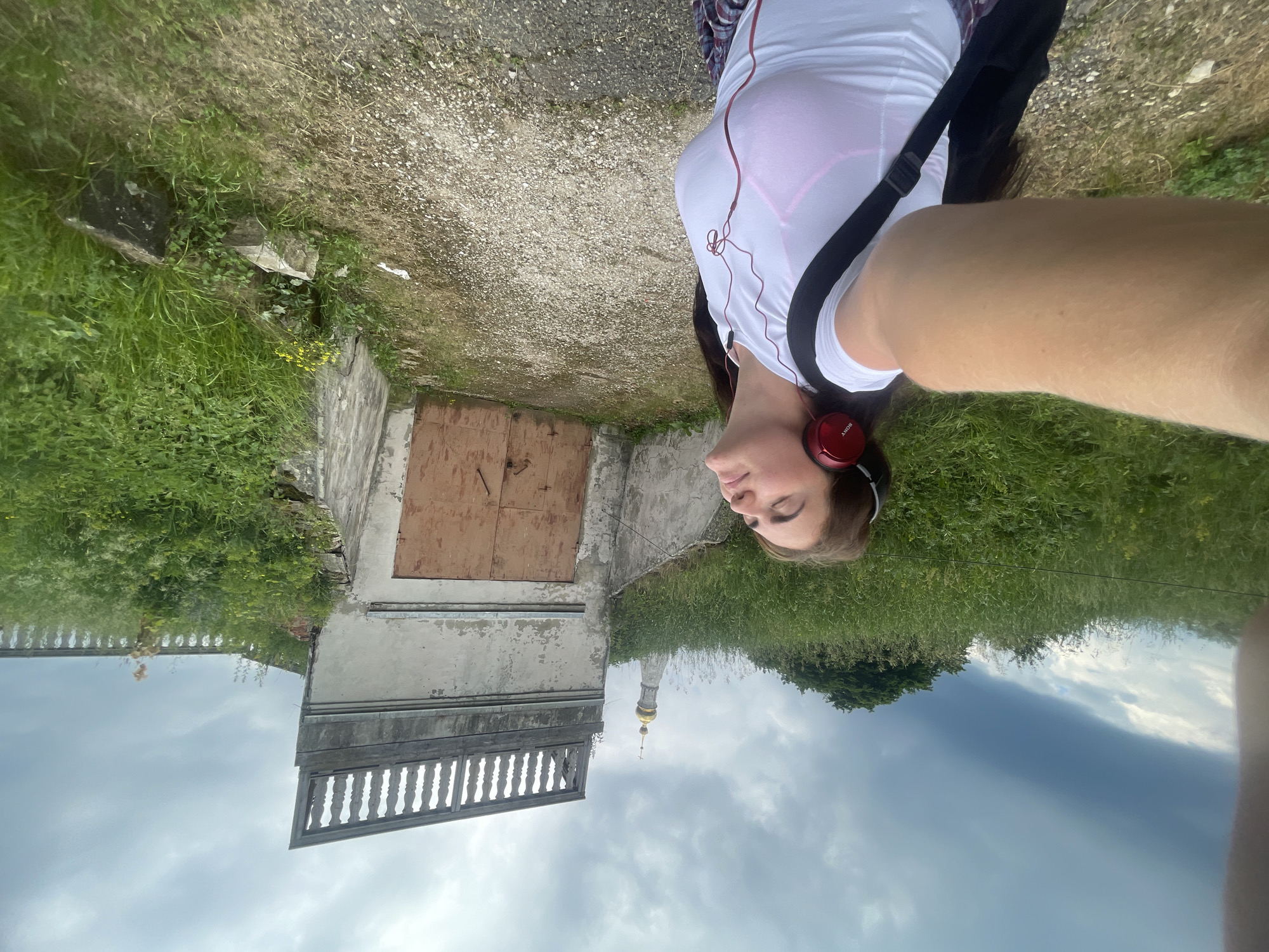 In Ostrov, Russia, on a cloudy day, a woman with dark hair is taking a selfie outdoors. She is wearing red headphones and a white t-shirt. The background features a dilapidated building with a large, weathered wooden door and a balustrade on top. Green grass and trees surround the area, and a golden-domed tower, likely part of a church, is visible in the distance. The ground is a gravel path.