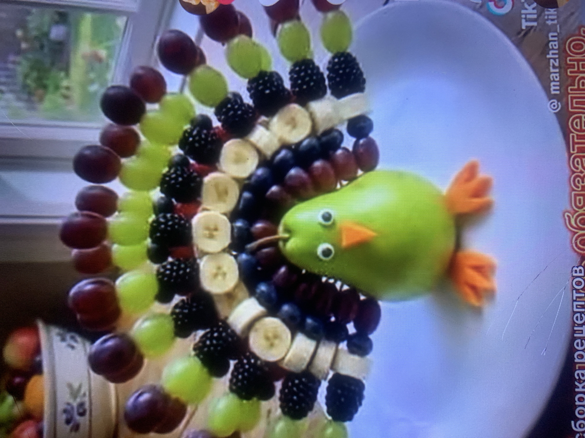 This media file is a close-up, slightly blurry image of a fruit platter arranged to resemble a bird. The bird's body is a bright green apple, with small black circles for eyes and a triangular orange piece for a beak. The bird is positioned on a white plate. Extending from the bird is a skewer laden with various fruits, forming what appears to be the bird's tail or body. These fruits include green grapes, dark red grapes, blackberries, and slices of banana. In the background, a window can be seen with greenery outside, suggesting an indoor setting with natural light. There is also a glimpse of a decorative basket to the left of the frame, filled with more fruit. On the right side of the image, overlaid text is visible, indicating the social media platform TikTok with a username "@marzhan_tik" and some text in Cyrillic script that appears to be "сборка рецептов" (recipe assembly). The overall impression is of a creative and healthy food presentation, likely for a celebration or as a visually appealing snack. The lighting suggests it might be daytime.
