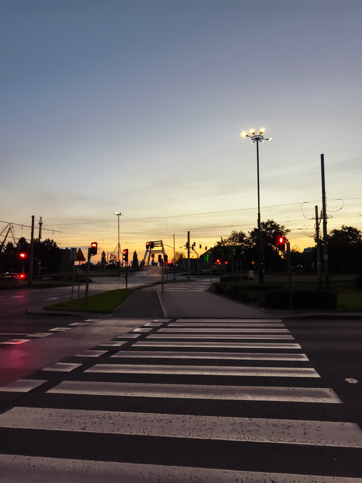 The image captures a street scene at dusk in Gdańsk, Poland, with a focus on a pedestrian crossing. The sky displays a gradient of colors, transitioning from a pale blue at the top to a warm yellow and orange hue near the horizon, indicating the setting sun. Several traffic lights are visible, most of them showing a red signal, suggesting that traffic is stopped. A bridge with an arched structure is visible in the background, illuminated by the twilight. Streetlights are also illuminated, providing additional light to the scene. The road surface is asphalt, and the pedestrian crossing is marked with white stripes. Trees and utility poles with overhead wires are present along the sides of the road. The overall atmosphere is one of quiet anticipation, possibly during a lull in traffic.