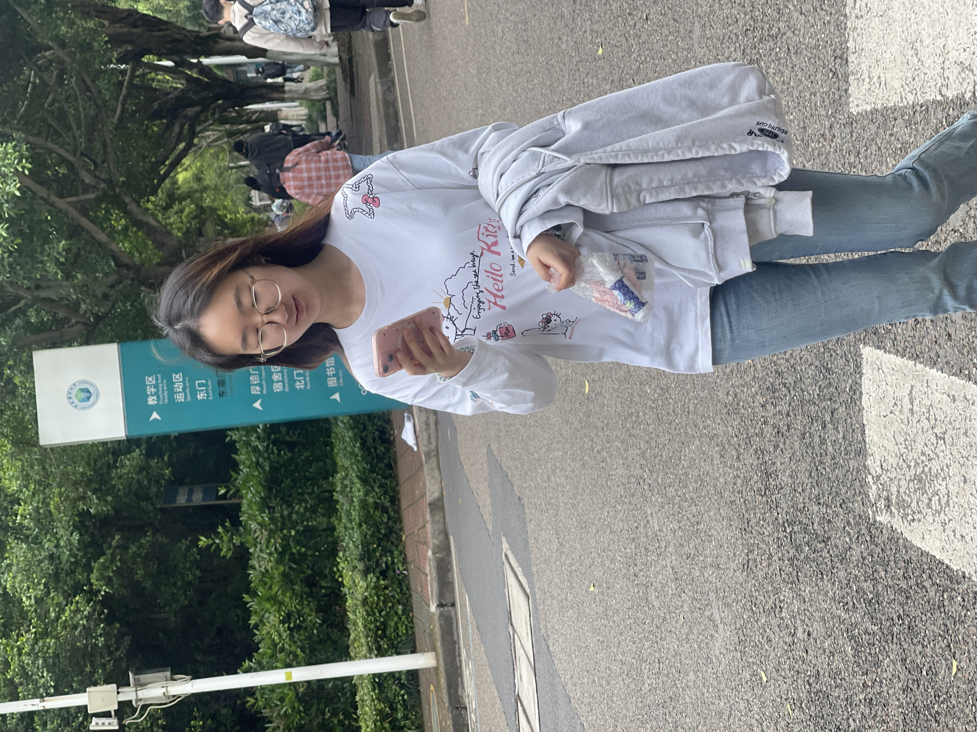 A young woman with long, dark hair and glasses is the central subject of this outdoor, eye-level shot. She is dressed in a white long-sleeve t-shirt with a colorful "Hello Kitty" graphic print and light blue jeans. Over her left arm, she carries a light gray hooded jacket. She is looking down at a pink smartphone held in her hands and also holds a small clear plastic bag containing what appears to be snacks or small items in her right hand.
The setting appears to be a university or campus environment in Chongqing High-tech Zone, China, characterized by a paved road, lush green trees, and a prominent blue-green directional sign. In the background, other blurred individuals can be seen walking amongst the trees. The lighting suggests it is daytime, likely sunny.
Visible text includes:
On the directional sign, in Chinese and English: "教学区 (Teaching And Study Area)", "运动区 (Sports Area)", "东门 (East Gate)", "厚德门", "宿舍区 (Dormitory Area)", "北门 (North Gate)", and "图书馆 (Library)".
On her t-shirt, text related to the Hello Kitty graphic: "Enjoying the sea breeze", "Hello Kitty", "Sand, sea & sun". Another visible text block on her clothing reads "1997 HEALING CAFE".