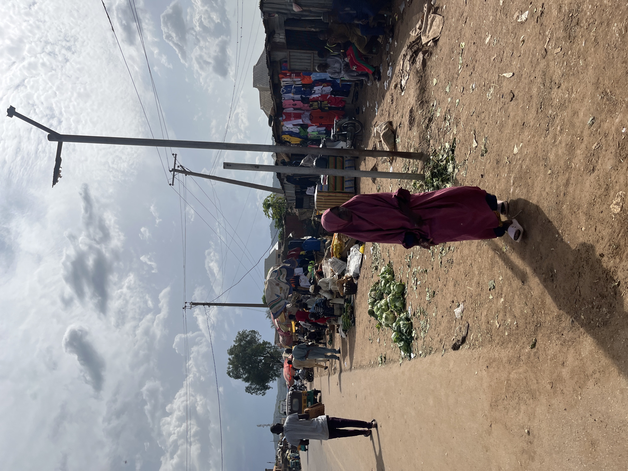 A street market scene is depicted in Jos, Nigeria, with a woman in a pink hijab and a long dress standing in the foreground, facing away from the camera. The market stretches along a dusty road, with various stalls and vendors visible. In the background, people are gathered, some tending to their stalls, while others appear to be customers. Cabbages and other produce are displayed on the ground. Clothing items hang from racks at one of the stalls. A motorcycle is parked near a stall. The environment appears somewhat cluttered, with debris scattered on the ground. The sky is cloudy, with sunlight filtering through.