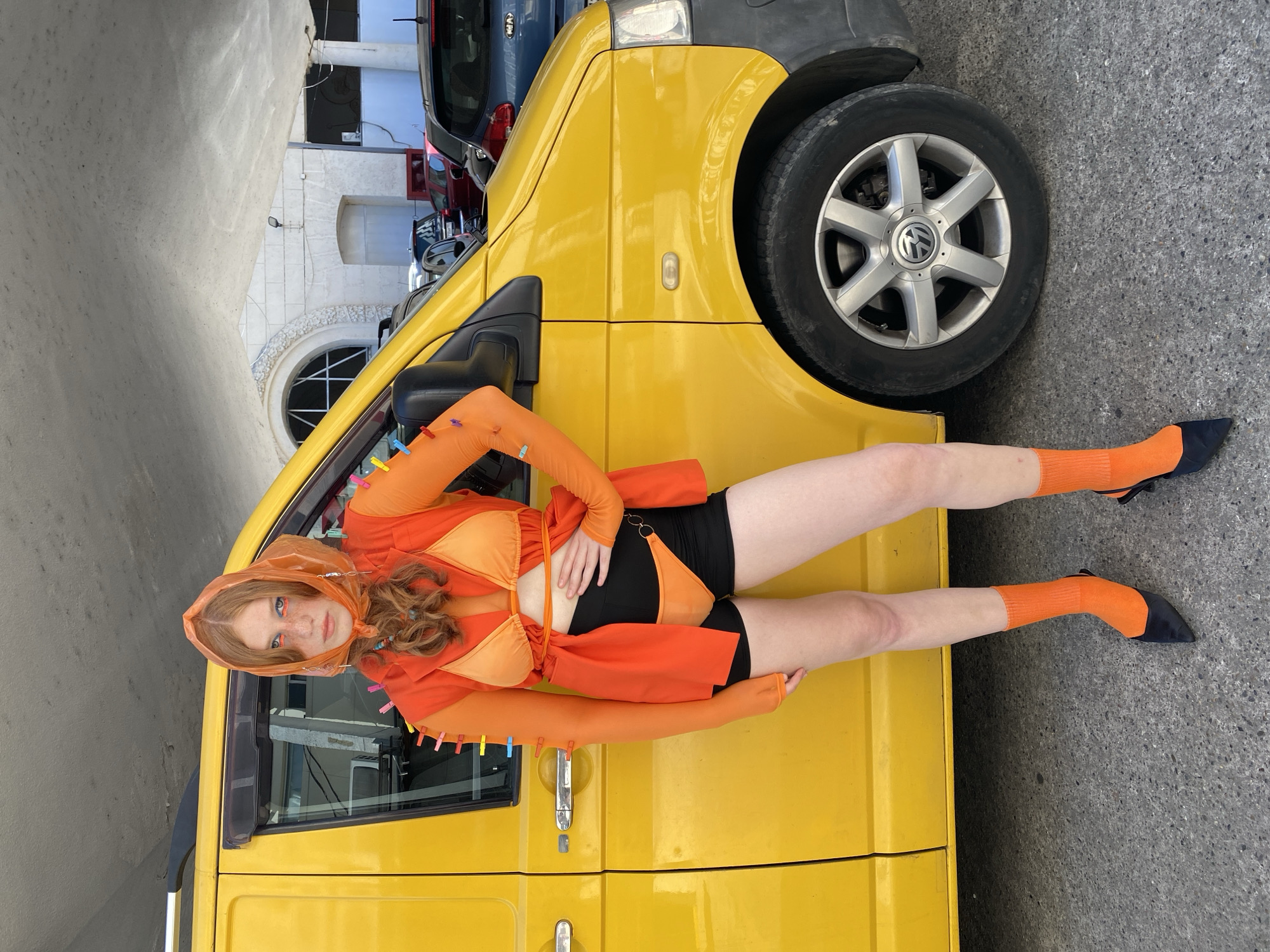 A young woman with long, wavy red hair and freckles is posing against the side of a bright yellow van. She is wearing an orange hooded top with detached sleeves, an orange bikini top, and black shorts. She also has on orange socks and black pointed-toe shoes. Her makeup includes orange eyeshadow and winged eyeliner. Clothes pins are attached to the edge of the van and her clothing. The woman is lying down with her legs extended, with one leg bent at the knee.
The van is parked on a gray asphalt surface, with a building visible in the background. The building appears to be made of light-colored stone and has arched windows. Other vehicles are parked behind the yellow van. The scene suggests it is daytime, with bright lighting. The location is identified as Sochi, Russia.