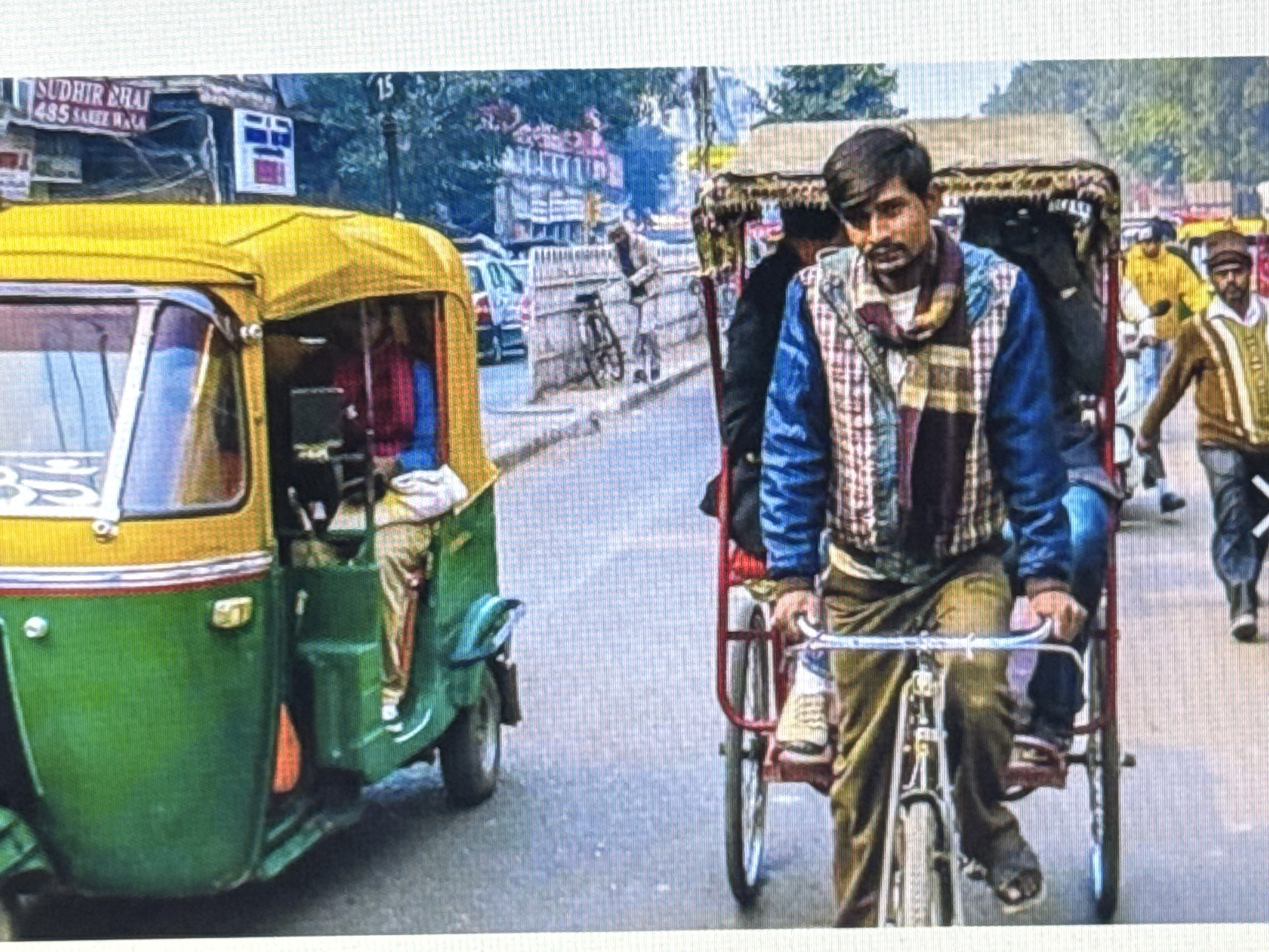 In the city of Unknown, Unknown, a street scene unfolds. On the left, a green and yellow auto-rickshaw is parked, its passenger door open, revealing glimpses of the interior. To the right, a man is pedaling a cycle rickshaw, his face looking directly at the camera. He wears a blue and plaid jacket with a scarf. The road is paved, and in the background, buildings and trees line the street. Other people and vehicles are visible further down the road, suggesting a bustling urban environment. A sign with "SUDHIR BHAI 435 SAREE WALA" is visible on a building in the background.