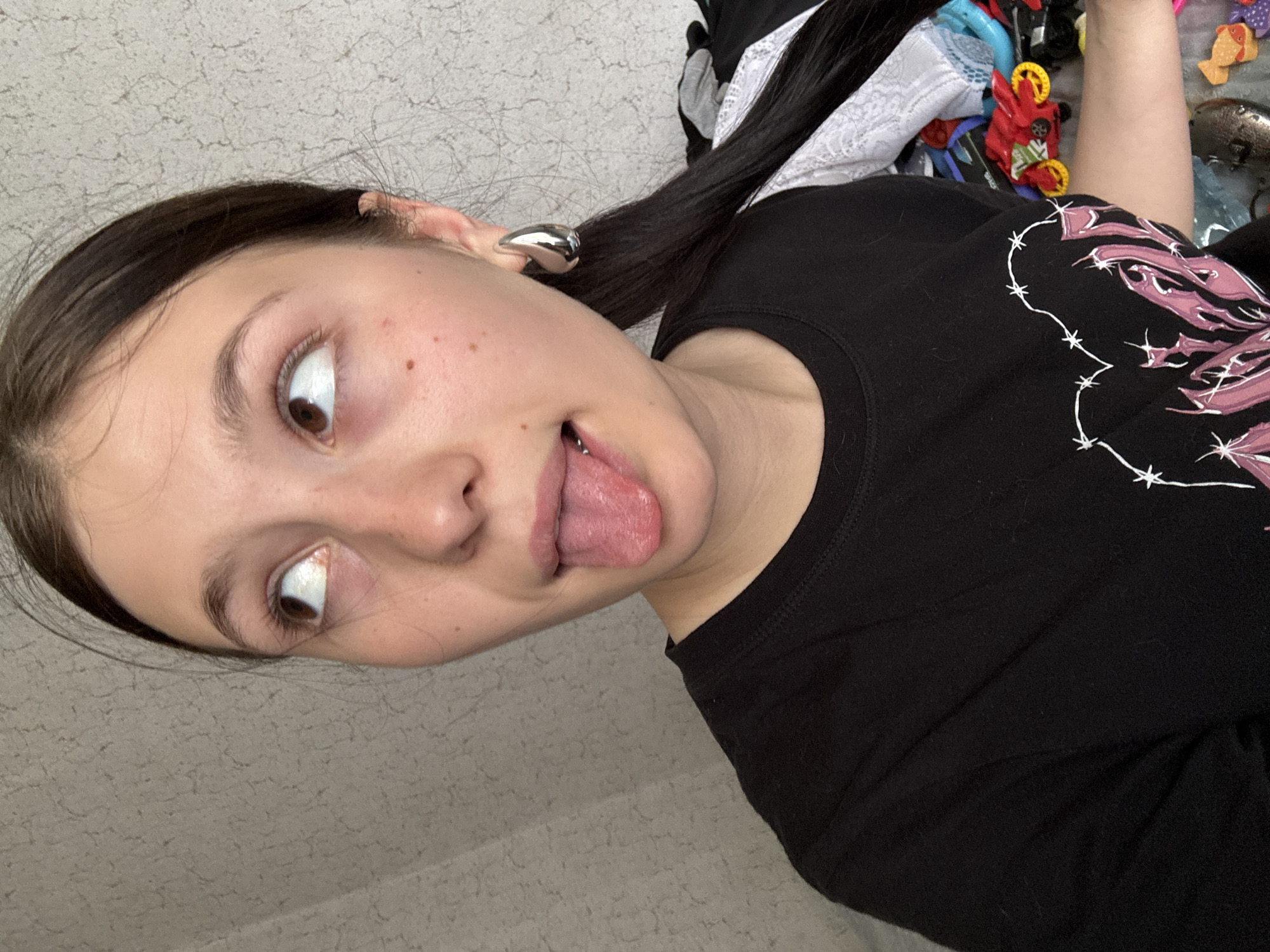 A young woman with dark hair is sticking her tongue out and looking to the side. She is wearing a black t-shirt with a white and pink design. She has an earring in her left ear. The background includes a gray wall and some toys and clothing on a surface behind her. There is no information about the city of Фоки, Russia in this image.
