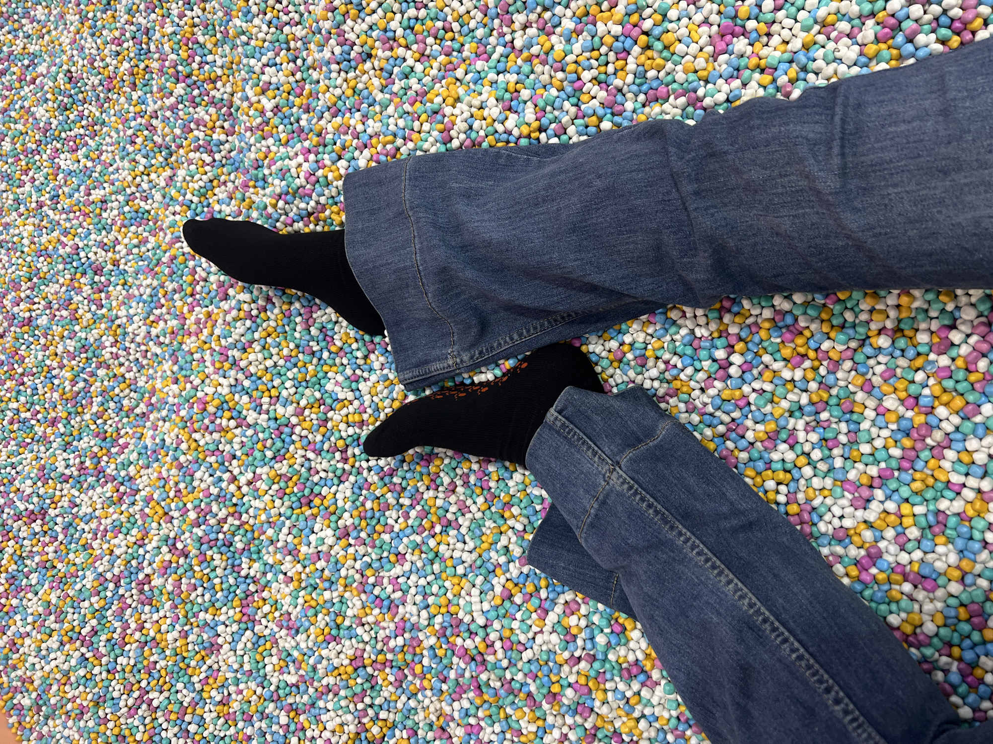 The image shows a close-up, overhead view of two pairs of legs submerged in a ball pit filled with small, colorful, pill-shaped objects. The objects are a mix of white, yellow, light blue, and pink. Both pairs of legs are wearing dark denim jeans, with one pair of legs also wearing black socks. The overall impression is a playful and sensory environment. The provided location of Kecamatan Pati, Indonesia, is a city on the island of Java.