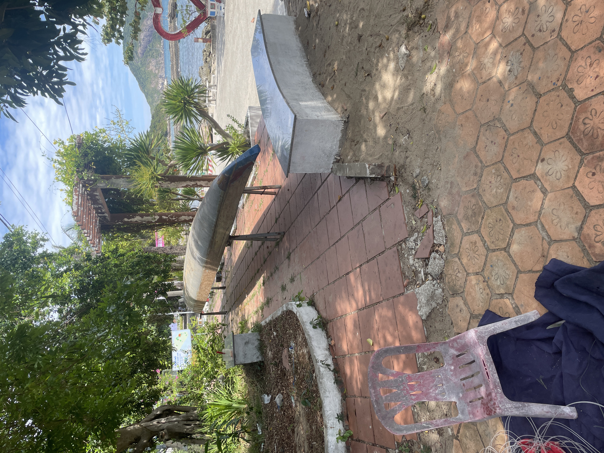 The image depicts an outdoor scene on a sunny day, likely in a coastal area. In the foreground, there is a weathered, pink plastic chair placed on a dark blue cloth, situated on a patterned hexagonal tiled walkway. Adjacent to the walkway is an area of sand with scattered debris. A long, narrow, metallic object, resembling a canoe or boat hull, is elevated on metal stands. Behind the boat, there's a curved, concrete bench with a polished stone top, bordering a sandy area. Further back, there's a garden bed with lush green plants and some scattered litter. A wooden pergola structure covered in vines is visible. The background features a body of water, a distant mountainous landscape, and a bright blue sky with scattered clouds. A large, red heart-shaped sculpture is also present near the water's edge, adorned with planters. The overall impression is one of a somewhat neglected public space, possibly a park or recreational area, near a beach. The specific location of Đà Nẵng, Vietnam, is not explicitly indicated within the image itself but may be inferred from external information.