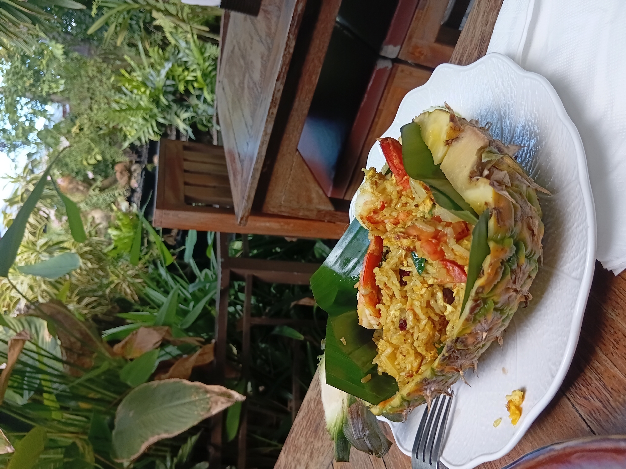 In Baan Lamai, Thailand, a plate of fried rice served inside a hollowed-out pineapple sits on a rustic wooden table. The rice is garnished with shrimp and green leaves, nestled within the pineapple's core. A fork rests on the white, leaf-shaped plate, next to a small pile of spilled rice. In the background, lush green foliage and part of a wooden chair suggest an outdoor dining setting, possibly a restaurant or cafe. The lighting indicates it is daytime.