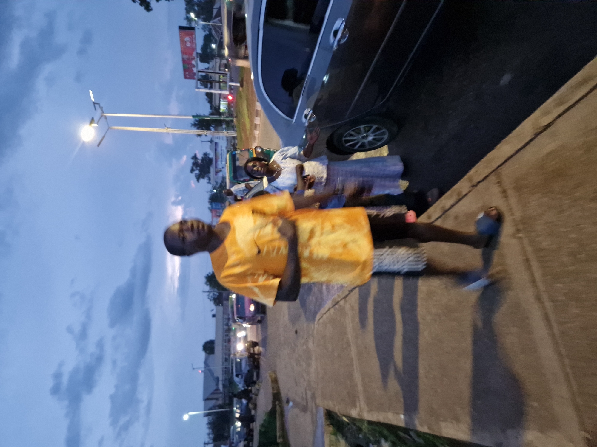 This is a full-body, medium shot, eye-level outdoor photograph taken at dusk on a sidewalk adjacent to a road in the city of Kabong, Nigeria. The sky is a deep blue with scattered, dark clouds. A street lamp illuminates the scene.
In the foreground, a man wearing a bright orange t-shirt with a tie-dye pattern and patterned shorts walks along the sidewalk. He is looking to his right. His feet are bare, and he is wearing blue flip-flops. He casts a long shadow on the sidewalk.
To the man's right, a woman in a light blue, patterned dress and matching headscarf stands near a dark gray car. She is looking slightly to her left and appears to be speaking to someone. Her left hand is resting on the car. Behind her, a green auto-rickshaw is visible.
The road behind them has several vehicles, including a red car, a motorcycle, and other cars. Further in the background, buildings and trees are visible, as well as more streetlights and advertisements. The overall impression is of a busy urban environment at twilight.