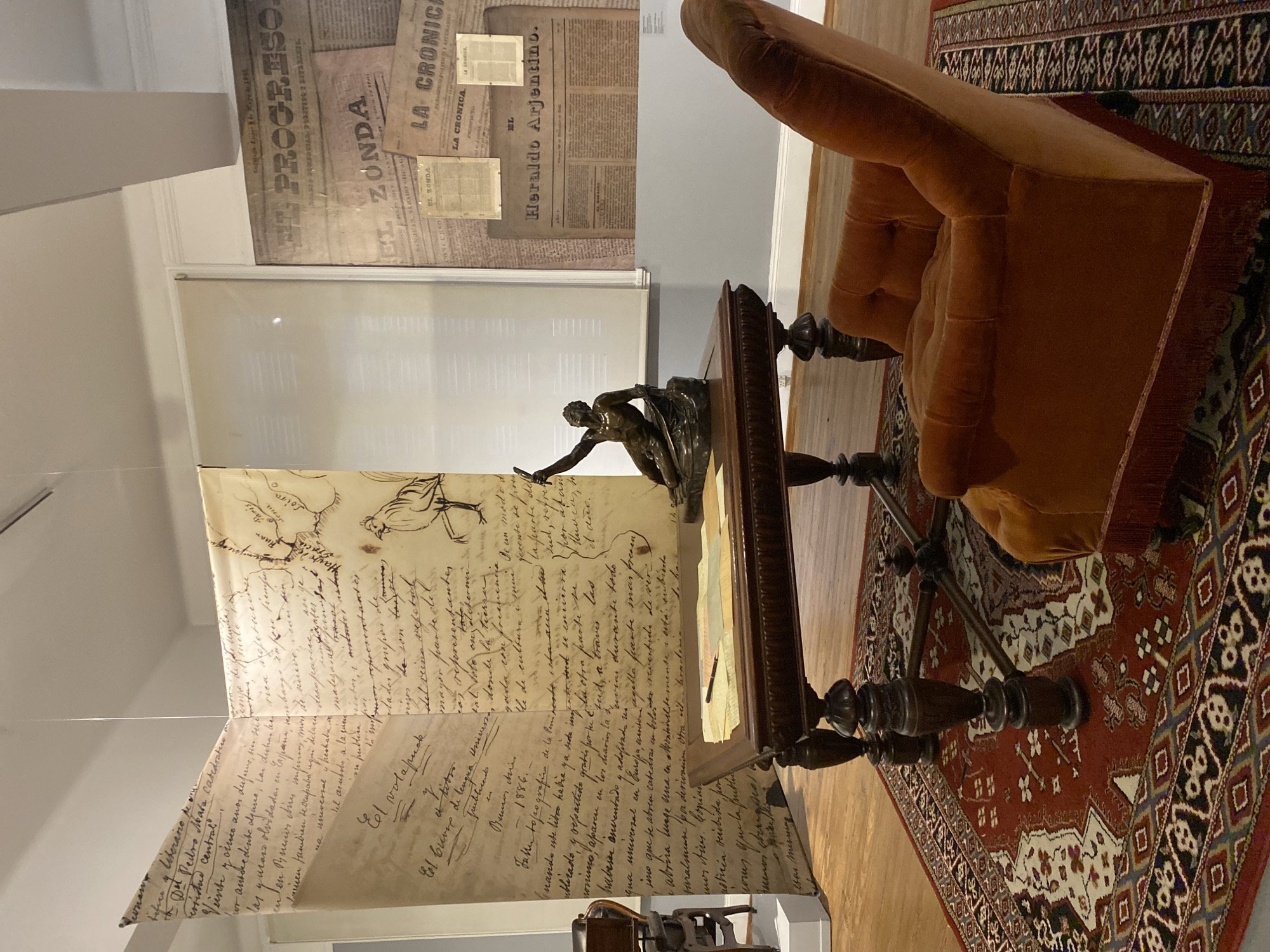 The image shows a room in what appears to be a museum or historical exhibition in Buenos Aires, Argentina. The room features several displays. On the wall behind a desk are newspaper clippings with headlines such as "EL PROGRESO," "EL ZONDA," "LA CRONICA," and "EL Heraldo Argentino." To the left of the desk, a large banner displays handwritten text in Spanish, adorned with a sketched map and a drawing of a bird.
A dark wooden desk is prominently positioned, topped with a bronze sculpture depicting a figure in action. To the right of the desk, there is an orange armchair, partially visible. A patterned rug covers the floor beneath the desk and chair.
The overall environment suggests an exhibit focused on the history of Buenos Aires, likely showcasing its newspapers and literary heritage.