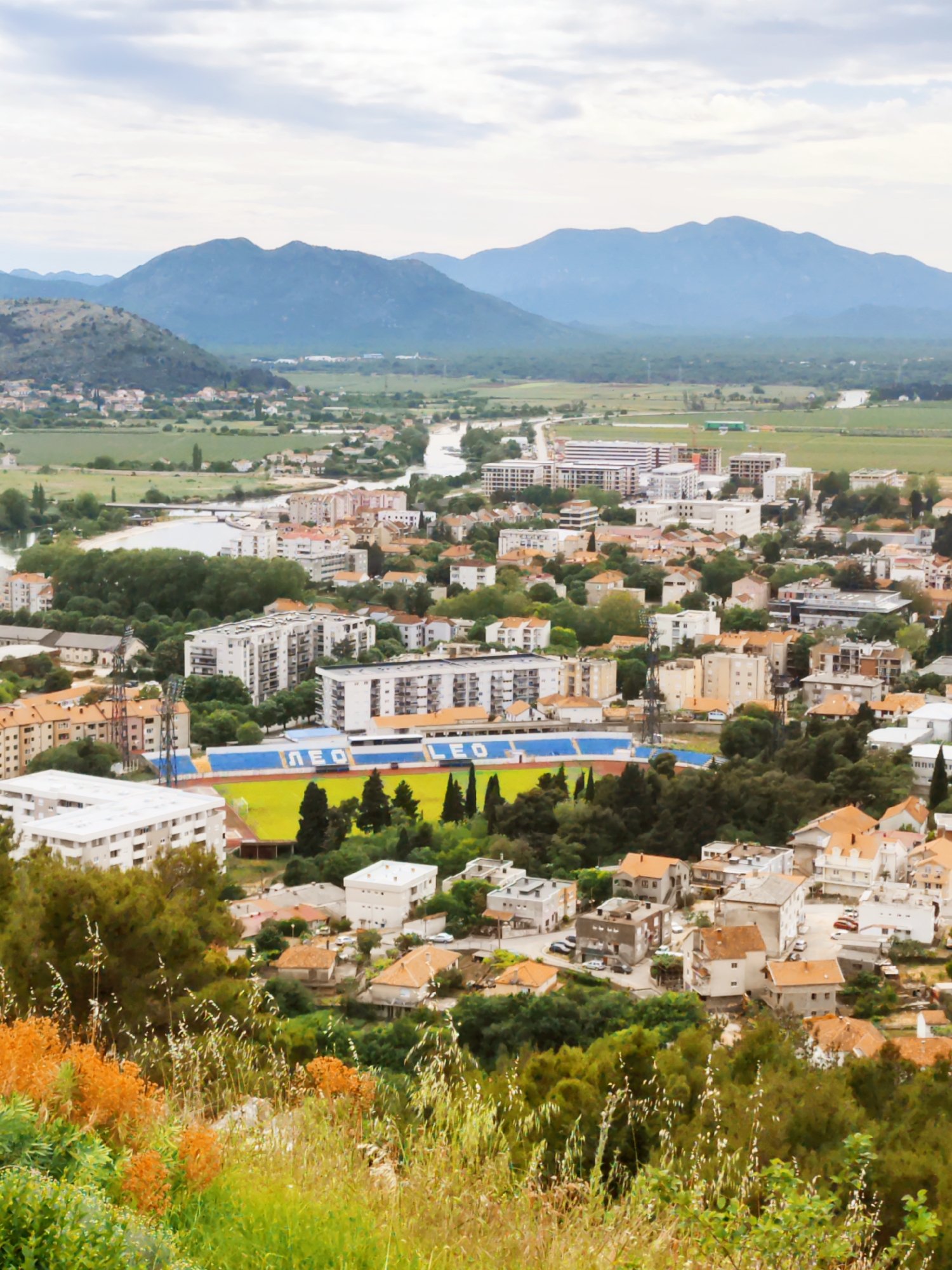 The image shows an aerial view of Trebinje, Bosnia and Herzegovina. A prominent feature is a football stadium with blue seats, and "LEO" is written in white letters. The city is built up with houses and buildings of various sizes and colors, many having orange roofs. Behind the city, there is a river winding through a green valley and mountains in the background under a cloudy sky. In the foreground, there is vegetation, including tall grass and bushes.
