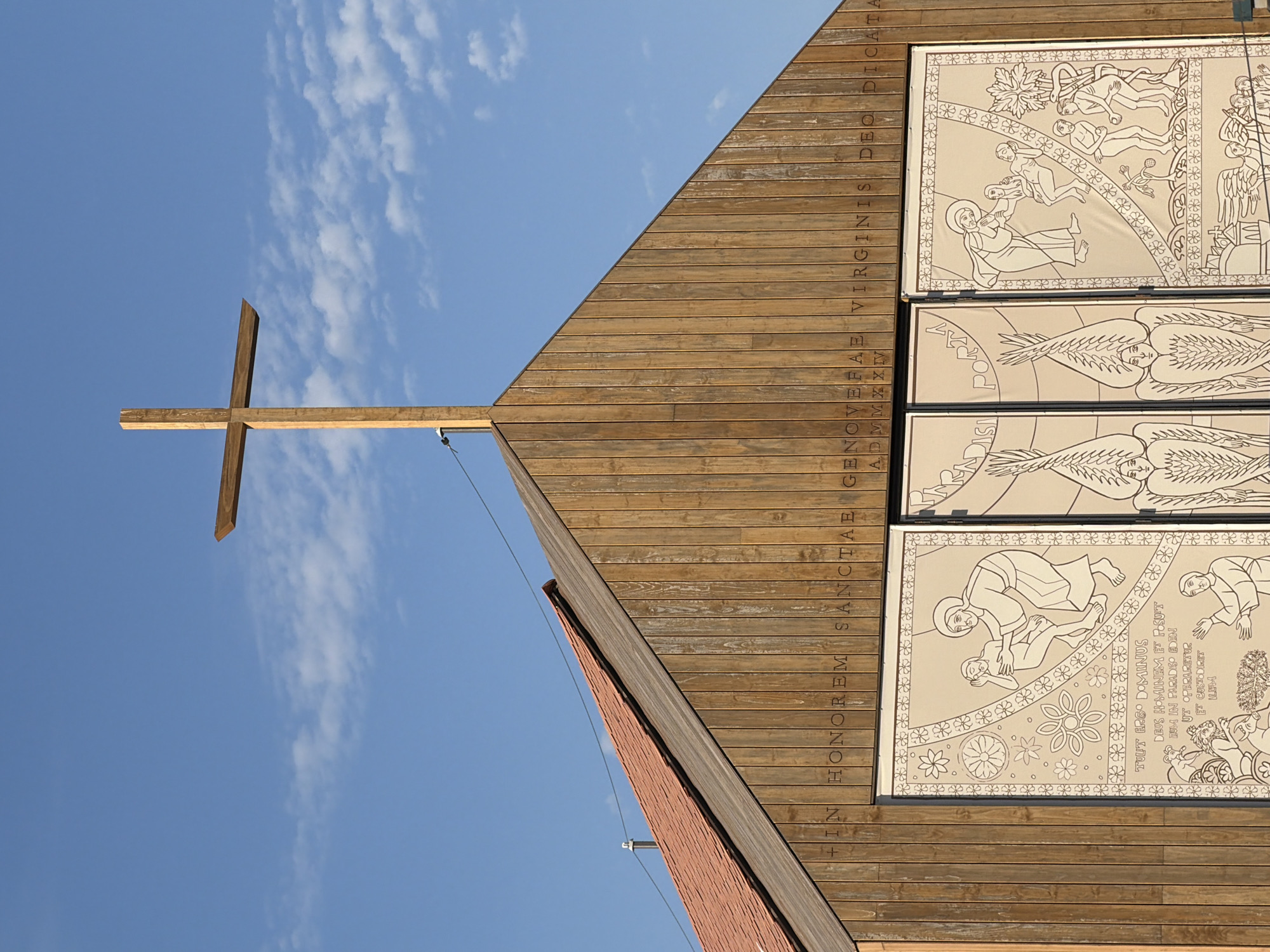The image shows the facade of a modern wooden church with a large wooden cross on top, set against a clear blue sky with scattered clouds. The church features large, decorated panels below the cross, depicting religious scenes in outline style. An inscription in Latin is visible on the wooden facade above the panels. The architecture is simple and contemporary. The church is identified as the Santuario de Santa Genoveva Torres Morales in Majadahonda, Spain.