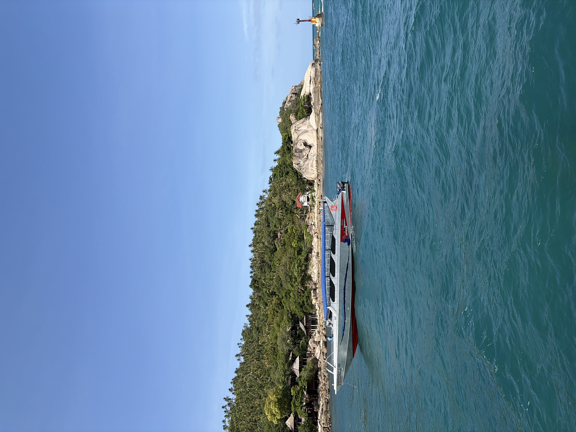 A high-angle, eye-level, medium shot captures a speedboat moving through turquoise waters, with a lush, tropical island and a lighthouse in the background under a clear blue sky. The speedboat, predominantly silver with red and blue accents, navigates the choppy sea, its engines visible at the stern. A lone figure stands on the deck of the boat. The island, a steep incline covered in dense green foliage and palm trees, is dotted with several small, traditional wooden structures with thatched roofs, suggesting a resort or village. Rocky outcrops punctuate the shoreline. To the right of the island, a red and yellow striped lighthouse stands on a rocky formation, warning of the shallow waters. The water itself exhibits gentle waves and a clear, vibrant blue-green hue, indicative of tropical waters.