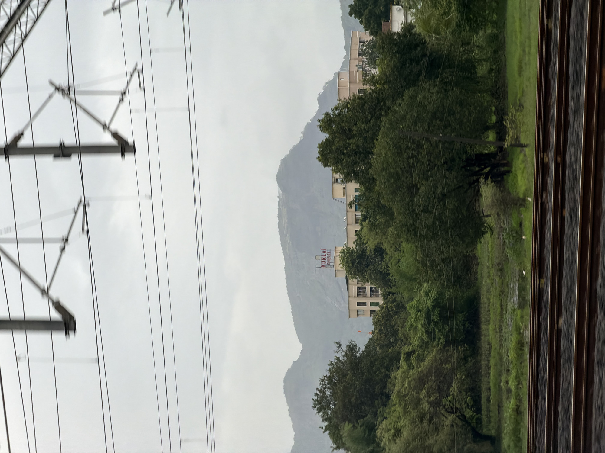 The image captures a view from a moving train in Saphale, India, with overhead railway electrification lines dominating the upper portion. Below the lines, a cluster of buildings, including a light-colored apartment complex with the name "KURLA COMPLEX" visible in red lettering on its rooftop, is nestled amongst lush green trees. In the background, rolling hills shrouded in mist or clouds suggest a slightly overcast day. The foreground features train tracks, indicating the perspective of someone traveling by rail. The environment appears to be a mix of urban development and natural landscape, characteristic of a semi-urban or suburban area.