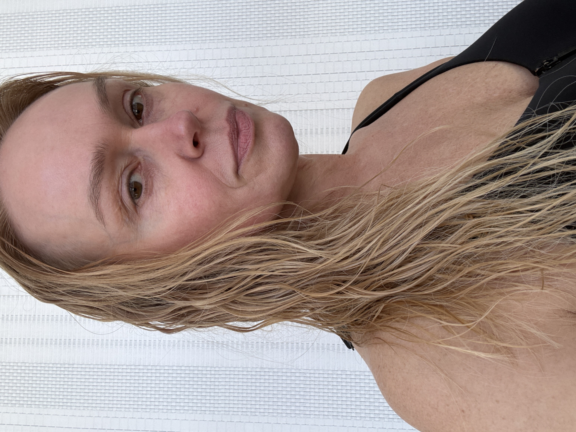 A woman with light brown hair is facing the camera. Her hair appears wet. She is wearing a black top with a strap visible on her shoulder. The background is a white textured surface. The location is not discernible from the image. The city of Chaikovsky, Russia, is not visible or implied.