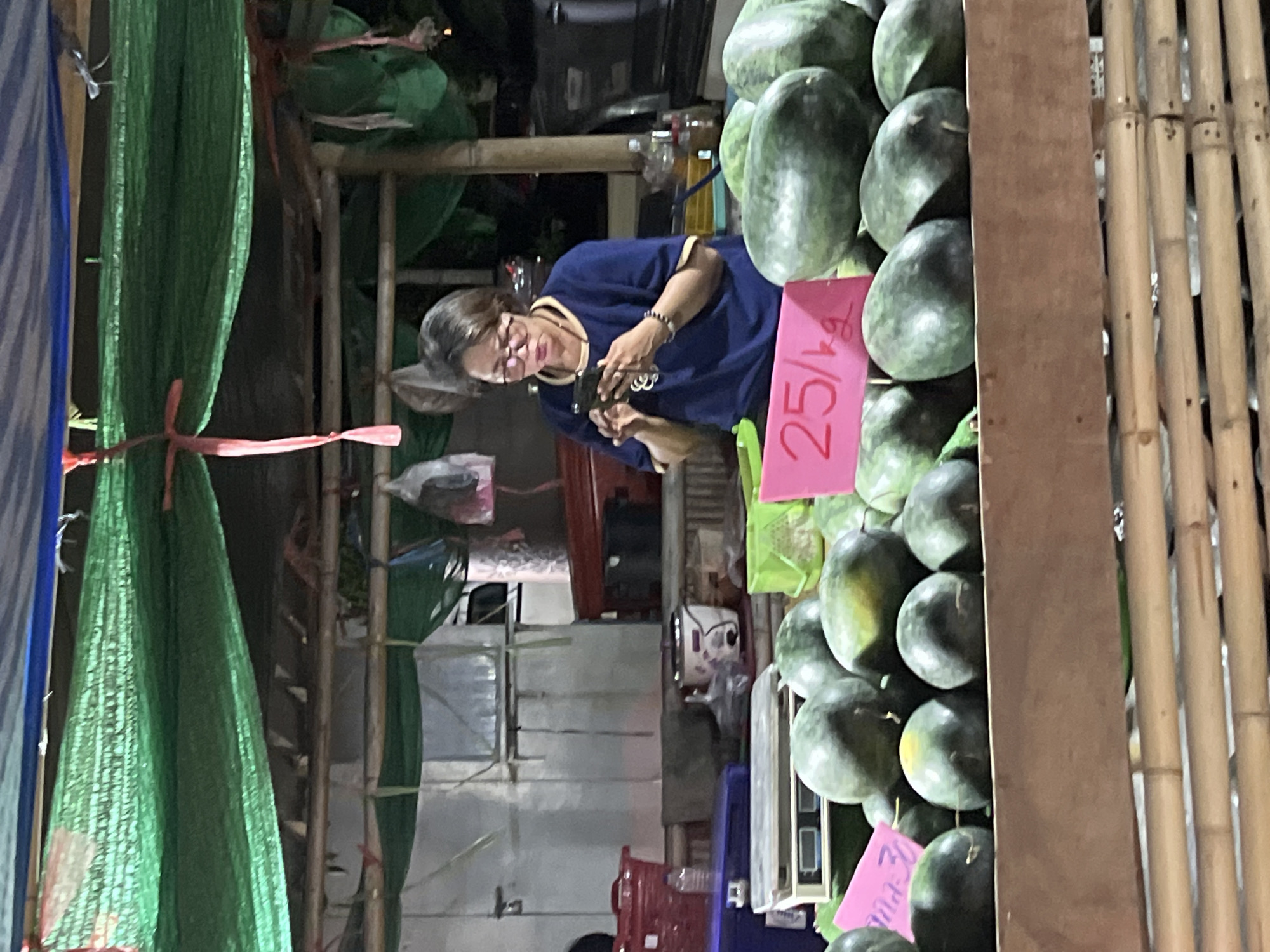 This image depicts a street vendor in Ko Pha-ngan District, Thailand, selling watermelons at a market stall. A woman, likely the vendor, is standing behind a display of watermelons and is looking at her mobile phone. She is wearing a blue t-shirt, glasses, and a necklace. The watermelons are arranged on wooden shelves and in baskets. A pink sign with "25/kg" written on it, indicating the price, is placed in front of the watermelons. The stall is covered with green netting, and bamboo structures are visible. The background shows various items typical of a market, including containers, bags, and what appears to be a rice cooker. The overall scene suggests a bustling market environment.