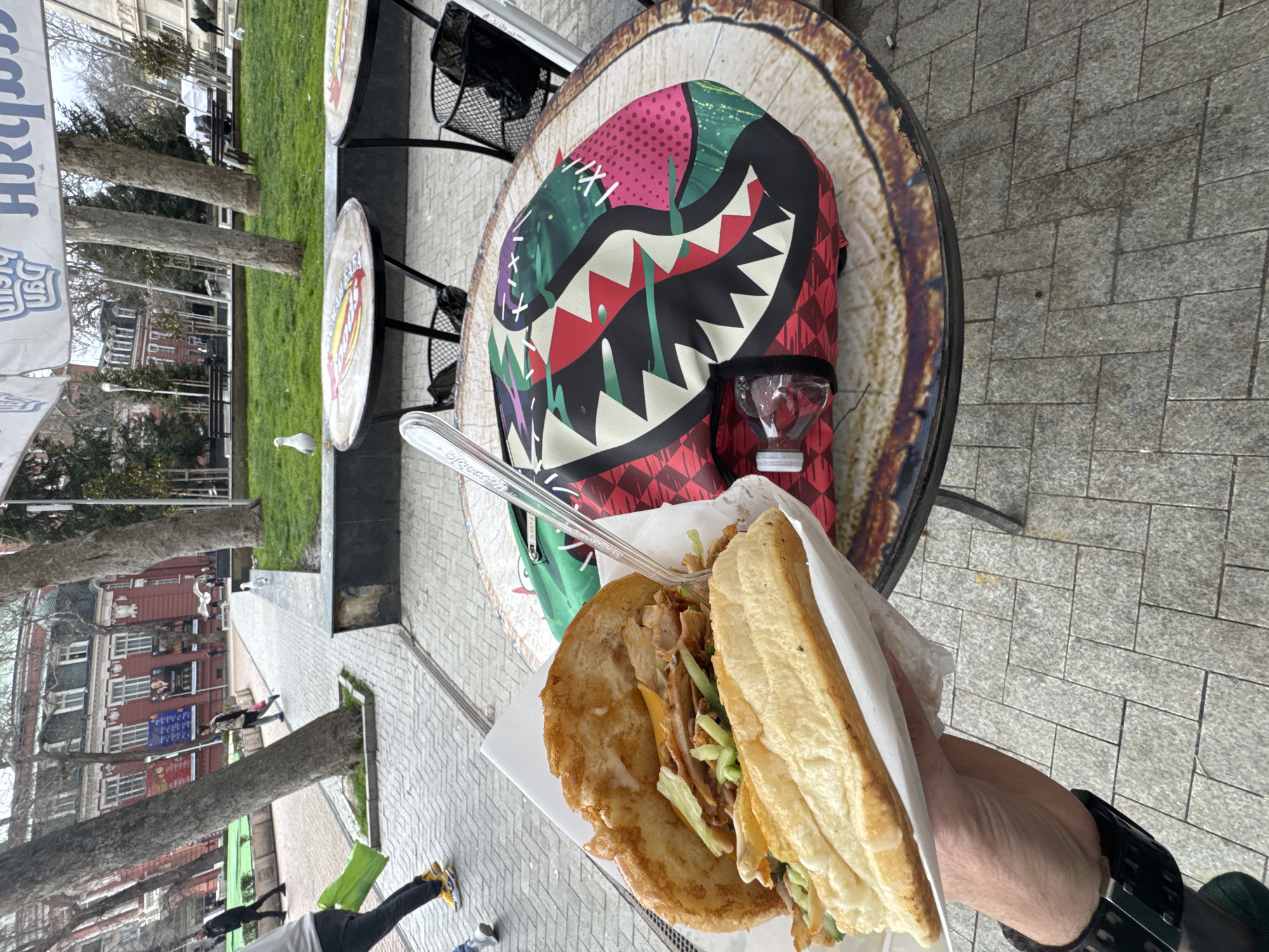 The image depicts a handheld sandwich against the backdrop of an outdoor scene, likely in Varna, Bulgaria. The sandwich, presented in white paper wrapping, is stuffed with meat and lettuce. It is held in a hand wearing a black watch. The backdrop includes a round wooden-patterned table with a colorful backpack placed upon it. A plastic fork rests atop the sandwich. Behind the table, there's a street scene featuring trees, buildings, and pedestrians. A white seagull is visible in the middle distance, and an awning with text in Cyrillic is partially visible in the top left corner. The urban environment includes bricked walkways.