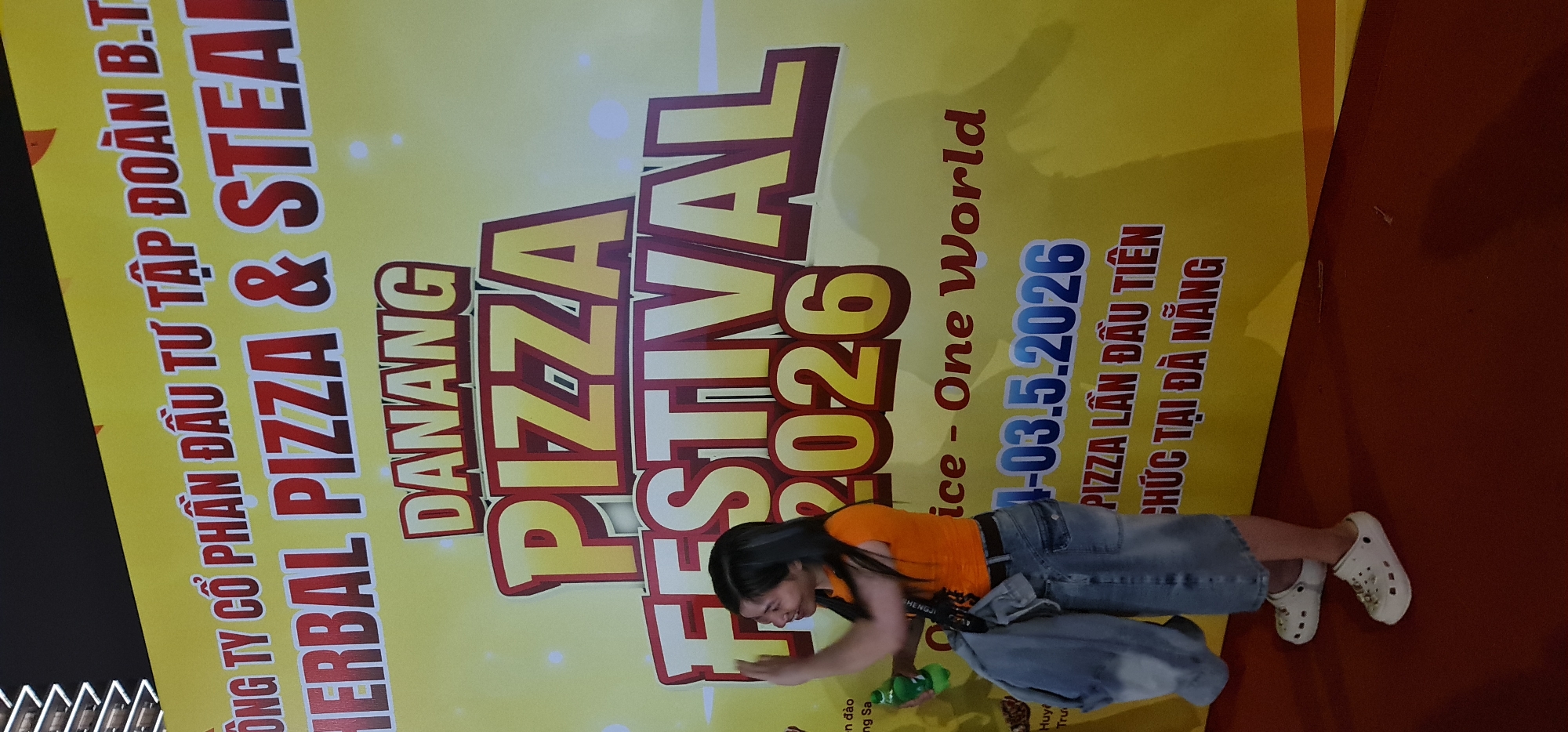A young woman is partially in the frame in front of a large, vertical advertisement for the "DANANG PIZZA FESTIVAL 2026" which will be held from 1-03.5.2026 in the city of Đà Nẵng, Vietnam. The advertisement also mentions "HERBAL PIZZA & STEAL" and the company "ÔNG TY CỔ PHẦN ĐẦU TƯ TẬP ĐOÀN B.T". The woman is wearing an orange shirt, blue jeans and white Crocs. She appears to be holding a green water bottle and gesturing towards the sign. A grey bag is draped over her side. The background is a red floor and what looks like a black wall.