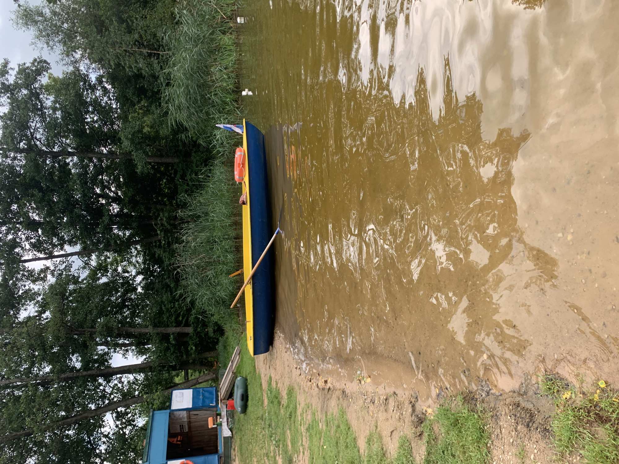 The image depicts a tranquil waterfront scene likely located in or near Грозаўскі сельскі Савет, Belarus. A blue and yellow rowboat rests by the sandy shore, its single oar partially submerged in the murky brown water. An orange life preserver and a small flag adorn the boat. Nearby, a green inflatable raft lies on the grass, and a small blue building with an open doorway stands on the left side of the frame, suggesting it might be a boathouse or storage facility. Tall trees and dense reeds line the far bank of the body of water, creating a natural backdrop. The water's surface reflects the overcast sky and the surrounding greenery. There are no people visible in the image.