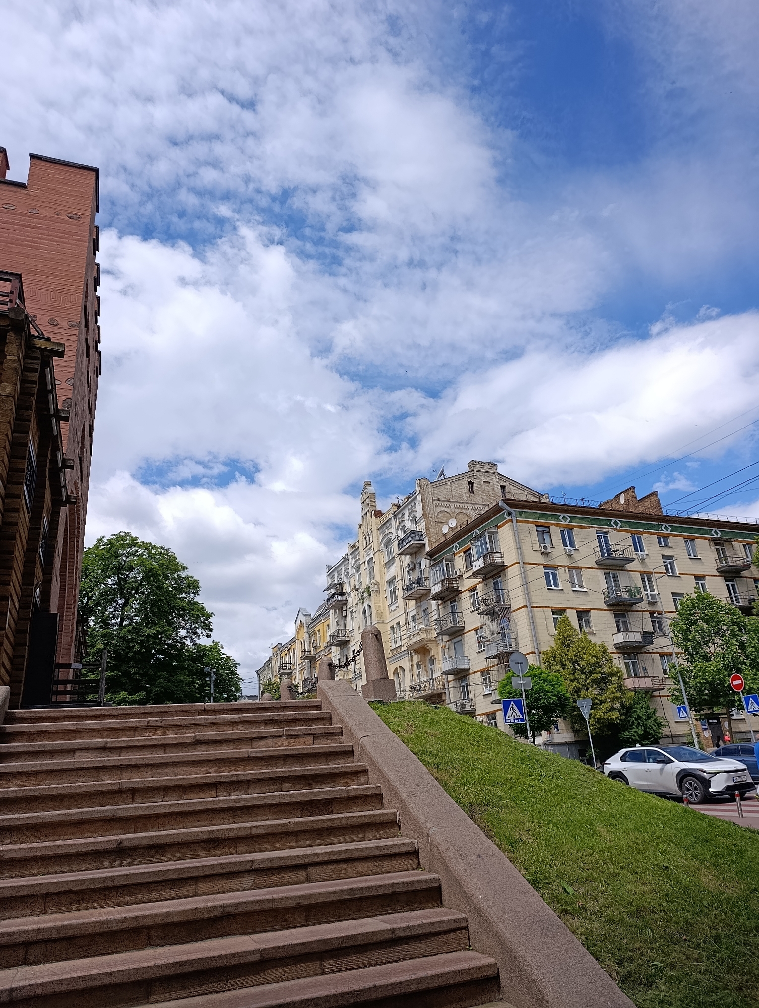 A wide-angle, low-angle outdoor shot captures a street scene in Kyiv, Ukraine, dominated by a large stone staircase in the foreground. The stairs are made of rough-hewn, reddish-brown stone with visible texture and wear. They ascend diagonally from the lower left towards the upper center of the frame, bordered by a granite railing. To the left of the staircase, a portion of a red brick building with decorative elements and windows is visible.
Beyond the staircase and to the right, the scene opens up to a street lined with buildings. A lush green lawn slopes upwards to the right of the stairs, dotted with trees and a white car. The buildings appear to be older, with multiple stories, balconies, and varied architectural styles, some painted in light colors and others with patterned facades. Trees are scattered throughout the scene, providing shade and greenery.
The sky above is a vibrant blue, filled with fluffy white cumulus clouds, suggesting a fair weather day. Several street signs and traffic signals are visible along the street. A few small birds are noted in the sky. The image's perspective emphasizes the scale of the staircase and the urban environment of Kyiv.