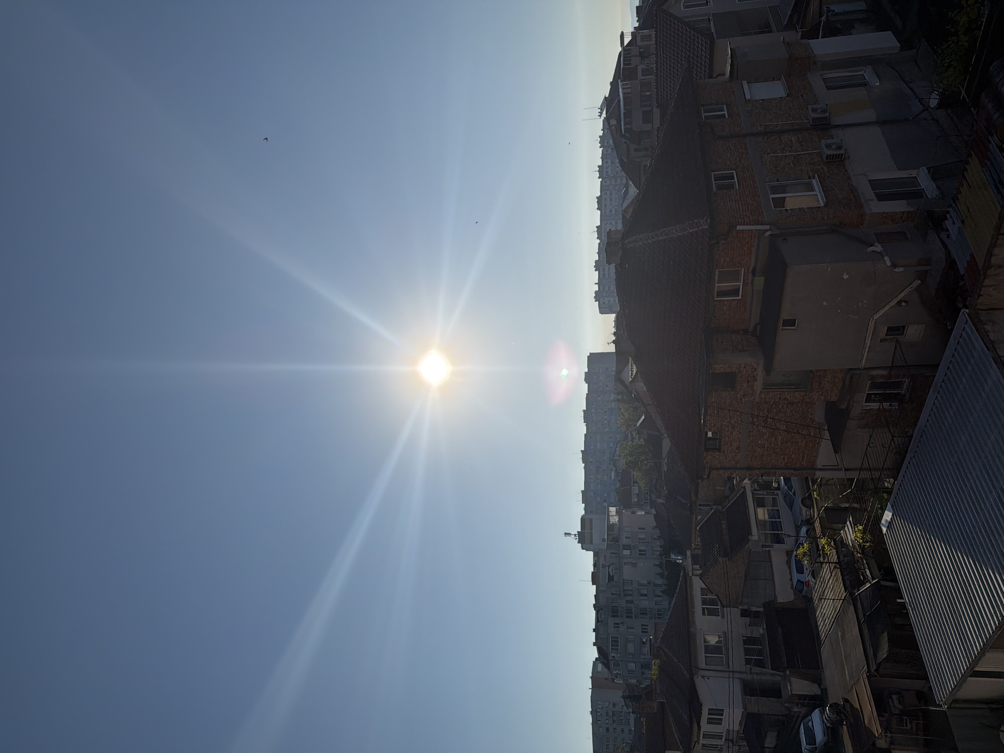 The image depicts an urban landscape under a bright, clear sky, likely taken from a high vantage point in Burgas, Bulgaria. The sun is prominently positioned in the sky, casting strong rays downwards. Buildings are visible, ranging from low-rise residential structures to multi-story apartment blocks. There is a rooftop visible in the lower right of the image. The rooftops are a mix of terra cotta tiled roofs and corrugated metal. There are a few birds in the sky. The light is so bright as to partially obscure detail.