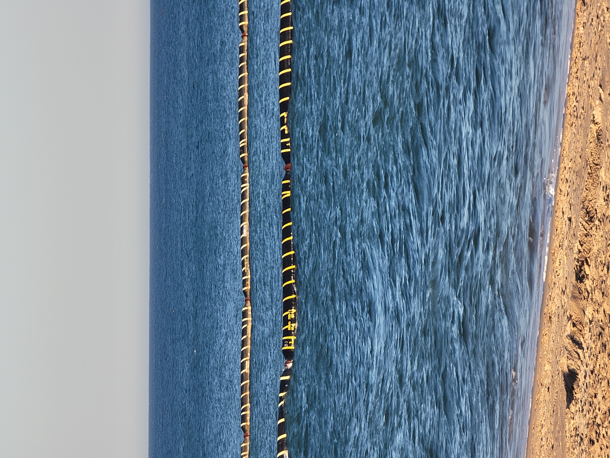The image shows a calm, blue sea with two parallel lines of yellow and black buoys floating on the water. The buoys are cylindrical and appear to be made of a flexible material. The sea is calm, with small ripples on the surface. The sky is clear and pale blue. The foreground of the image is a sandy beach. The scene is likely Quarteira, Portugal, as evidenced by the characteristic blue waters and sandy beaches of the region.
