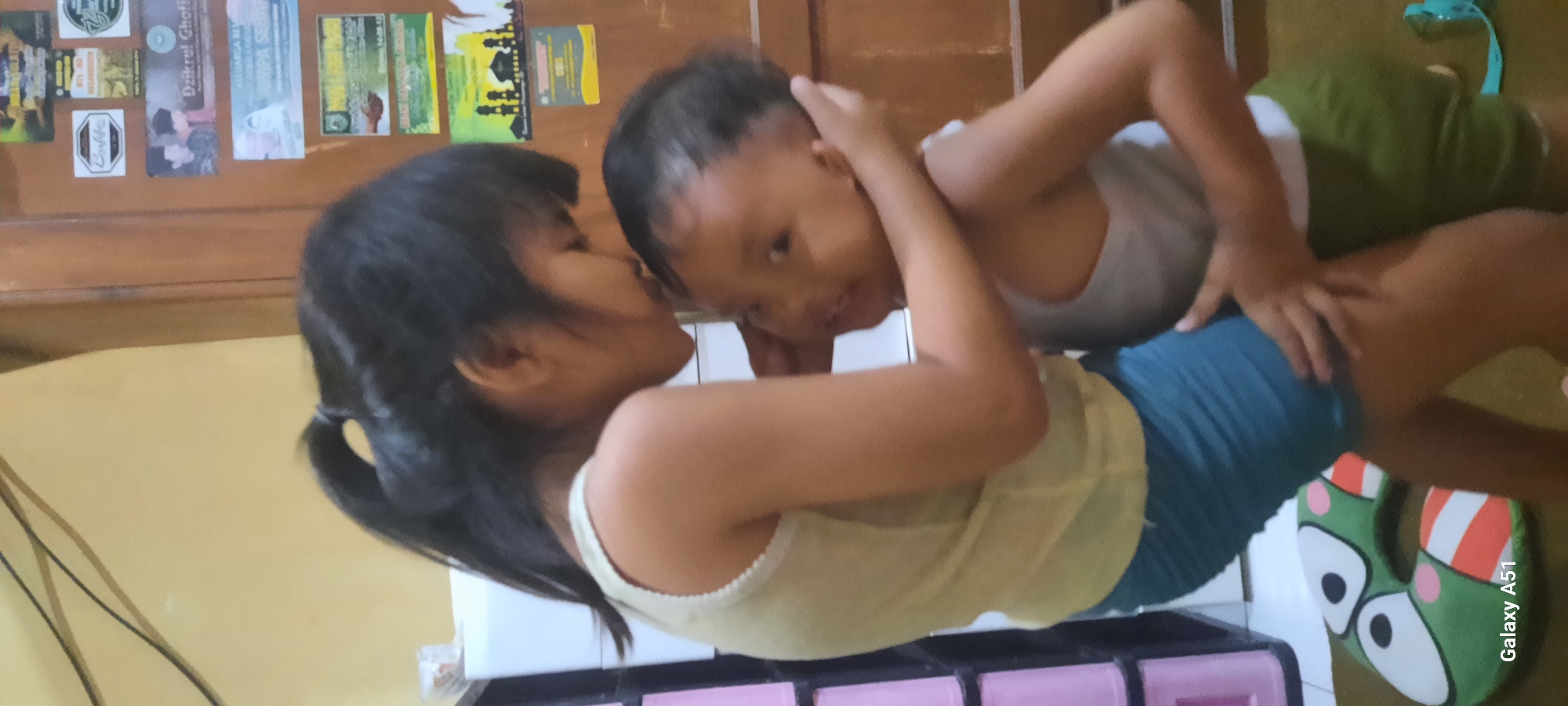 In the city of Jatitengah, Indonesia, two young children are engaged in an affectionate embrace. The older child, a girl with her hair tied back in a ponytail, is leaning forward and kissing the forehead of the younger child, a boy. Both children are wearing casual tank tops and shorts. The older girl has on a light yellow tank top and blue shorts, while the younger boy is wearing a grey tank top and green shorts. They are standing on a floor, possibly in a home environment, as indicated by the wooden door and various posters or papers affixed to it in the background. A white storage unit is partially visible to the left, and a colorful, cartoon-like pillow is on the floor near the children's feet. The lighting suggests an indoor setting, possibly during the day. The overall scene conveys a sense of sibling affection and warmth.