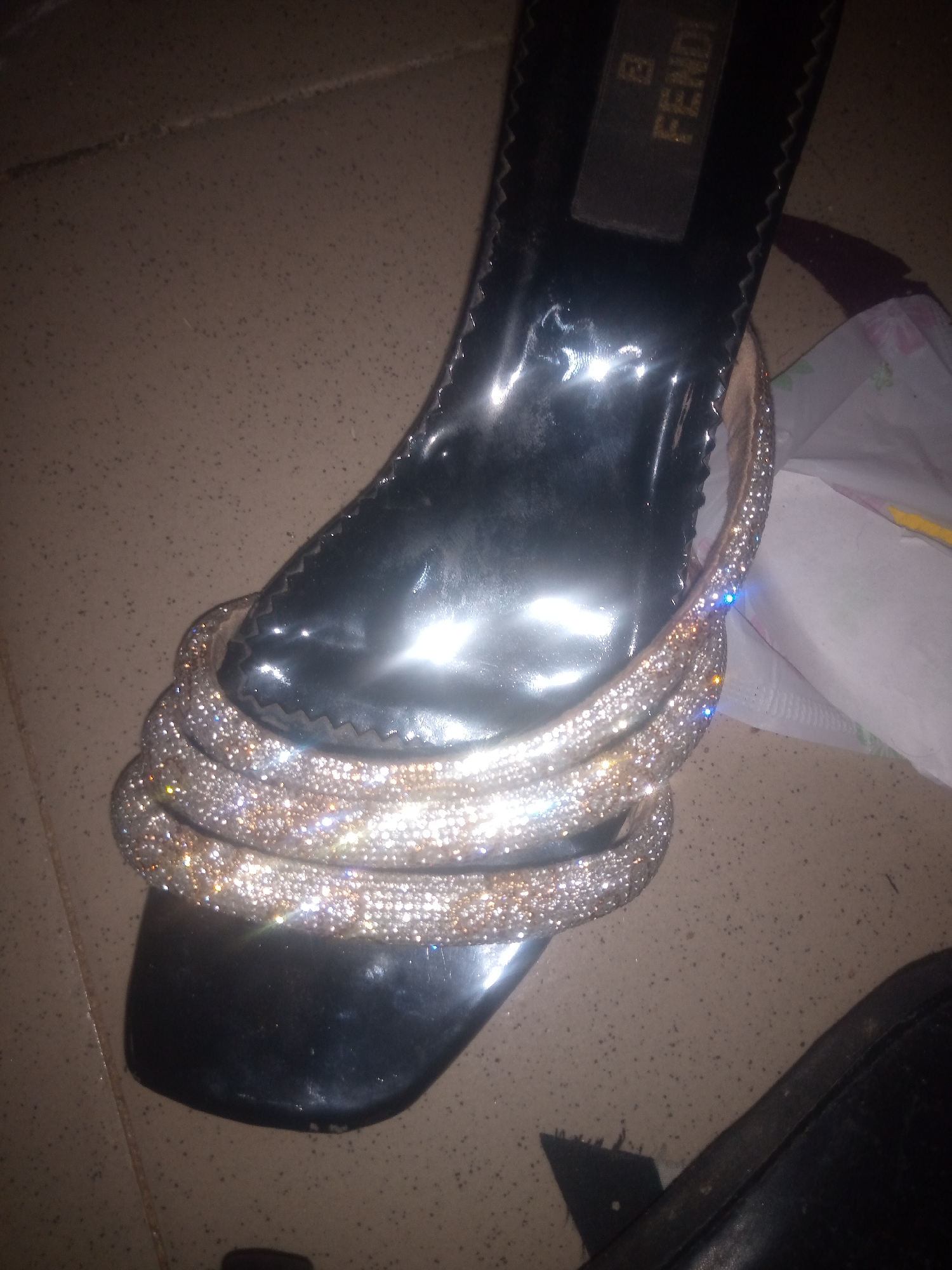 A close-up, high-angle shot shows a black Fendi sandal with multiple bands of rhinestones. The sandal is on a speckled tiled floor. To the right of the sandal, a white paper product with some colorful markings is visible. The lighting is dim, creating reflections on the sandal's surface. The image appears to be taken indoors. There are no people or animals visible in the frame.