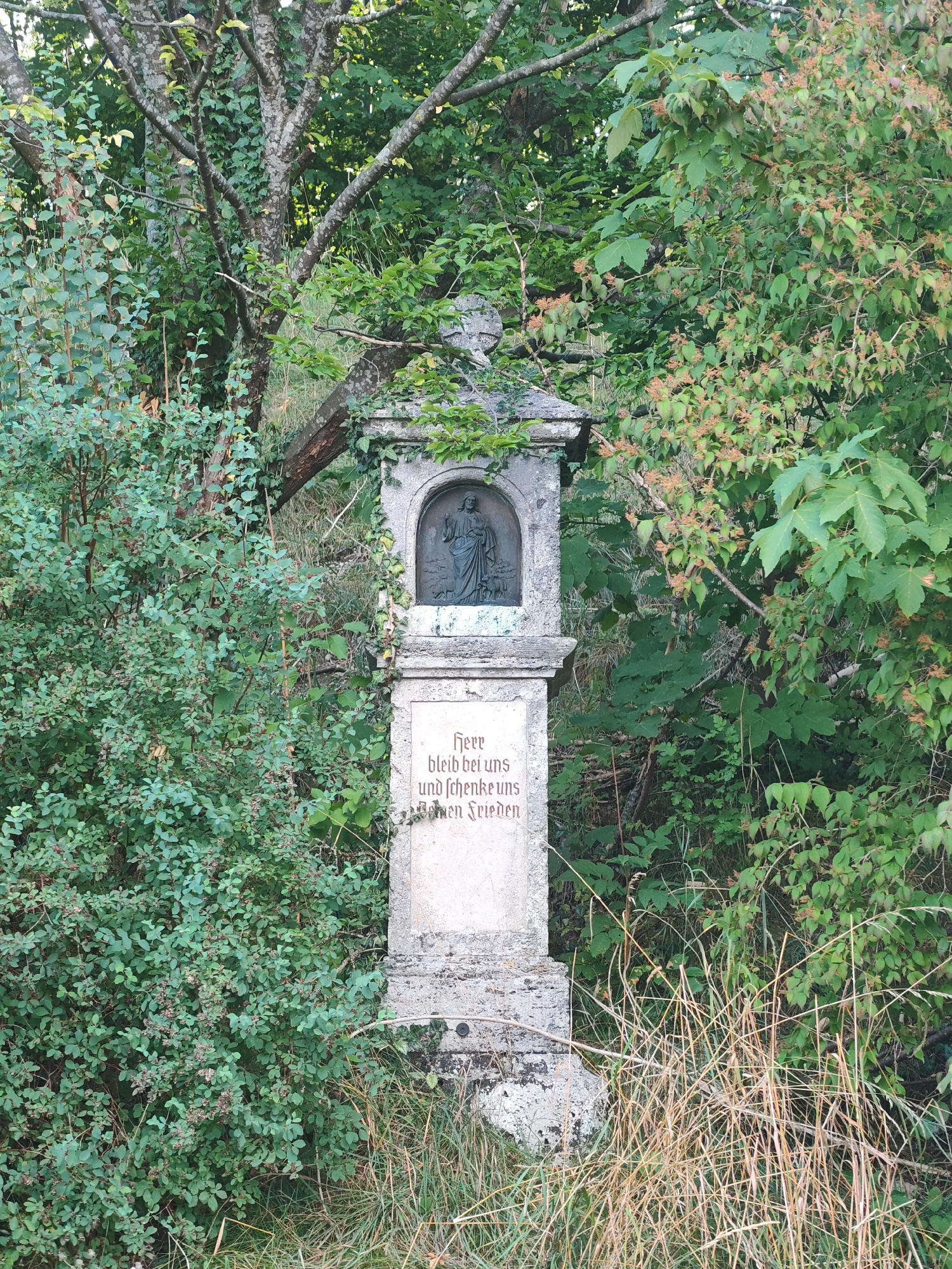 A weathered stone wayside shrine stands amidst dense foliage and tall grasses. The shrine is rectangular with a decorative top, featuring a relief carving of a religious figure in an arched niche. Below the niche, an inscription in Gothic script reads "Herr bleib bei uns und schenk uns deinen Frieden" which translates to "Lord, stay with us and give us your peace." The shrine is located in Obergünzburg, Germany, surrounded by overgrown vegetation, suggesting it is in a rural or natural setting. The lighting indicates it is daytime, with sunlight filtering through the trees, creating dappled shadows.