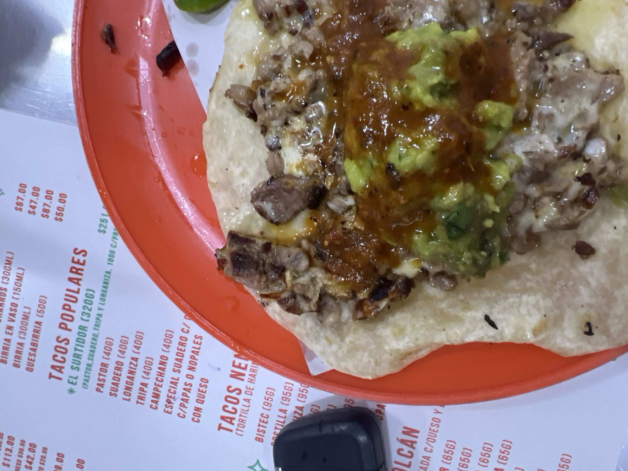 A close-up shot captures a single, generously topped taco resting on a bright orange plastic plate. The taco, on what appears to be a corn tortilla, is laden with chopped meat, possibly some melted white cheese, a reddish-brown salsa, and a prominent dollop of green guacamole or avocado salsa. Small, dark food remnants are scattered on the plate and around the taco.
The plate is positioned over a white menu, which is partially visible and displays text in Spanish with prices. The menu indicates a casual taqueria setting, typical of Mexico City, Mexico. Visible menu items include "TACOS POPULARES" (Popular Tacos) with varieties such as "PASTOR," "SUADERO," "LONGANIZA," "TRIPA," and "CAMPECHANO," each listed with a 40-gram portion size. There's also "*EL SURTIDOR" (The Assortment), which combines pastor, suadero, tripa, and longaniza. Other sections mention "QUESABIRRIA," "BIRRIA EN VASO," and "TACOS NEU" (likely "TACOS NUEVOS" or "New Tacos") made with "TORTILLA DE HARINA" (flour tortilla) and "BISTEC" (steak). Prices are displayed using the "$" symbol, consistent with Mexican pesos, ranging from approximately $20.00 to $251.00. A small black rectangular object, possibly a condiment holder, is visible in the foreground at the bottom.