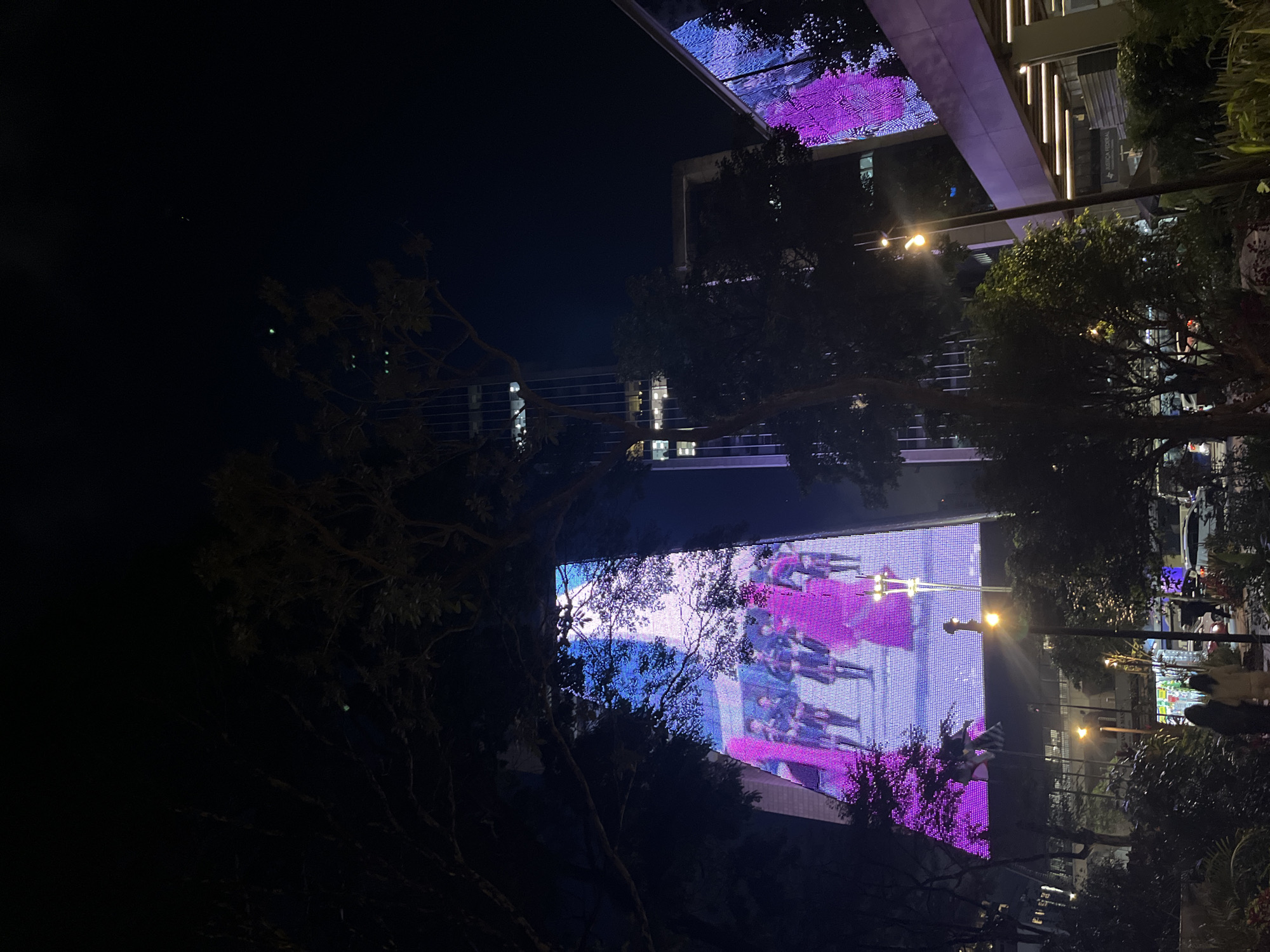 Captured at night, the image presents a street scene in what appears to be an urban environment. A building with a large video screen is visible, displaying vivid pink and purple hues. The screen's reflection illuminates surrounding trees. Streetlights cast a yellow glow on the foliage and the street below, where indistinct figures suggest pedestrian activity. The overall ambiance conveys a blend of natural and artificial light sources in a city setting.