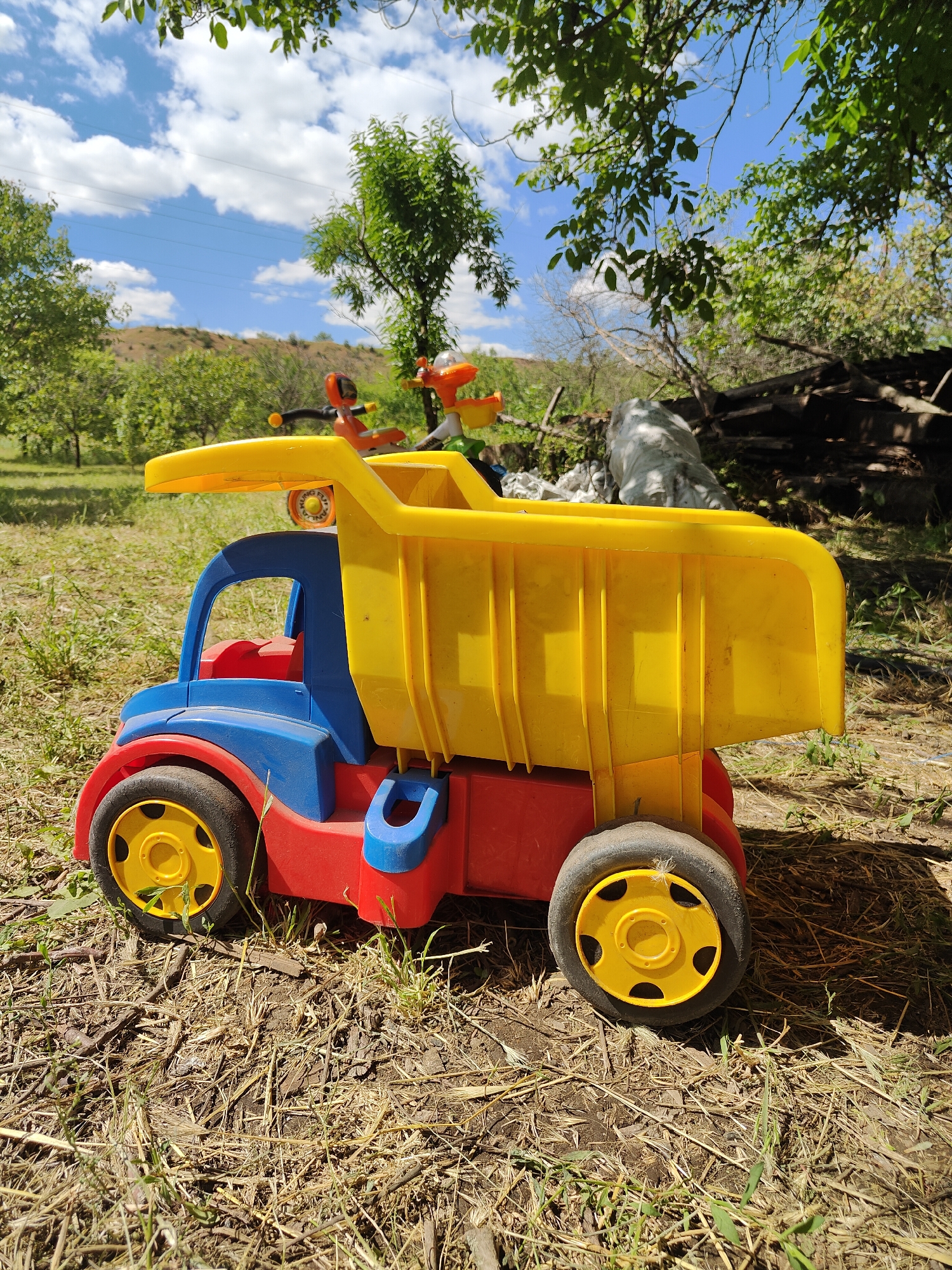 A vibrant yellow and red toy dump truck sits on dry, straw-covered ground in an outdoor setting. The truck is blue and red, with a large yellow dump bed. Two smaller toy figures, one orange and one yellow, are positioned on top of the dump truck's cab. In the background, there are green trees and a blue sky with fluffy white clouds, suggesting a sunny day. The ground is covered with dry grass, twigs, and dirt. The scene appears to be a backyard or a natural area. The time of day is likely daytime, given the bright sunlight casting shadows. There are no discernible emotions expressed as the subjects are toys and the scene is static. No visible text is present on the toy or in the background. The location context is Kryvyi Rih, Ukraine.