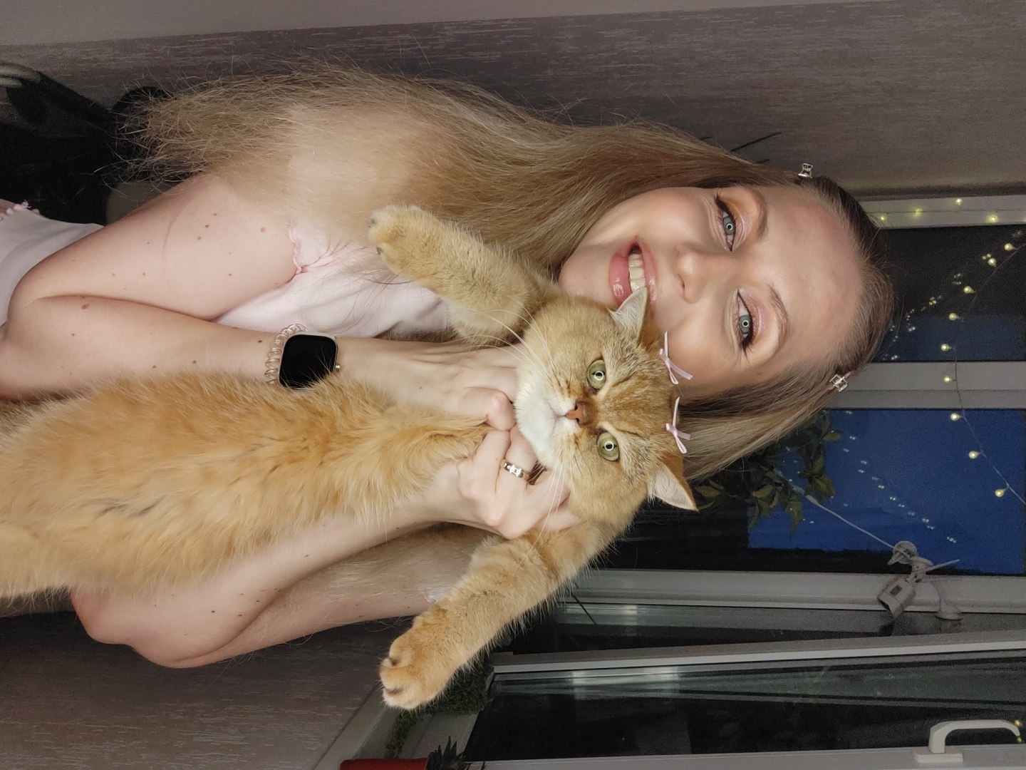 A woman, with blonde hair and pink eyeshadow, smiles broadly while holding a ginger cat. The cat has two small pink bows on its head. The woman is wearing a light pink top and a watch on her left wrist. The background features a window with string lights and a glimpse of a city skyline at night, possibly Krasnodar, Russia. The overall scene appears to be an indoor setting, perhaps a balcony or near a window in an apartment.