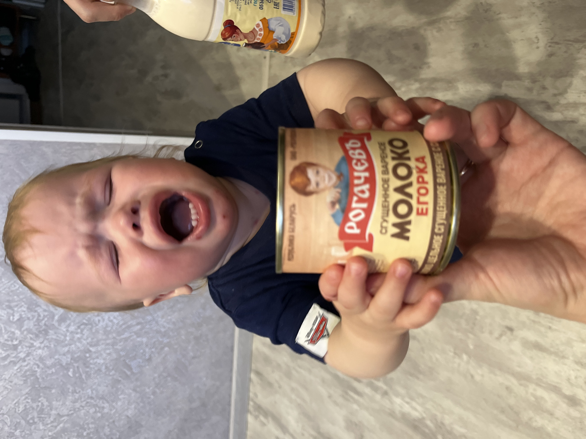 The image captures an indoor scene in Chaikovsky, Russia, centered on a distressed toddler and an adult hand. A light-haired child, estimated to be between one and two years old, is shown mid-cry with eyes tightly closed, mouth wide open displaying lower teeth, and face contorted in distress. The child wears a navy blue long-sleeved top with a "Cars" logo patch on the left sleeve. Both the child's small hands and an adult's fair-skinned hand, adorned with a gold-colored ring on the fourth finger, are holding a can of "РОГАЧЕВЪ СГУЩЕННОЕ ВАРЕНОЕ МОЛОКО" (Rogachev Boiled Condensed Milk), which features a boy on its label. To the blurred right background, a partial view of a white plastic bottle, likely containing "Золотая" (Zolotaya) condensed milk with a label depicting a woman in traditional attire, is visible. The background consists of a light-colored, textured wall and a similarly light, subtly patterned floor.
